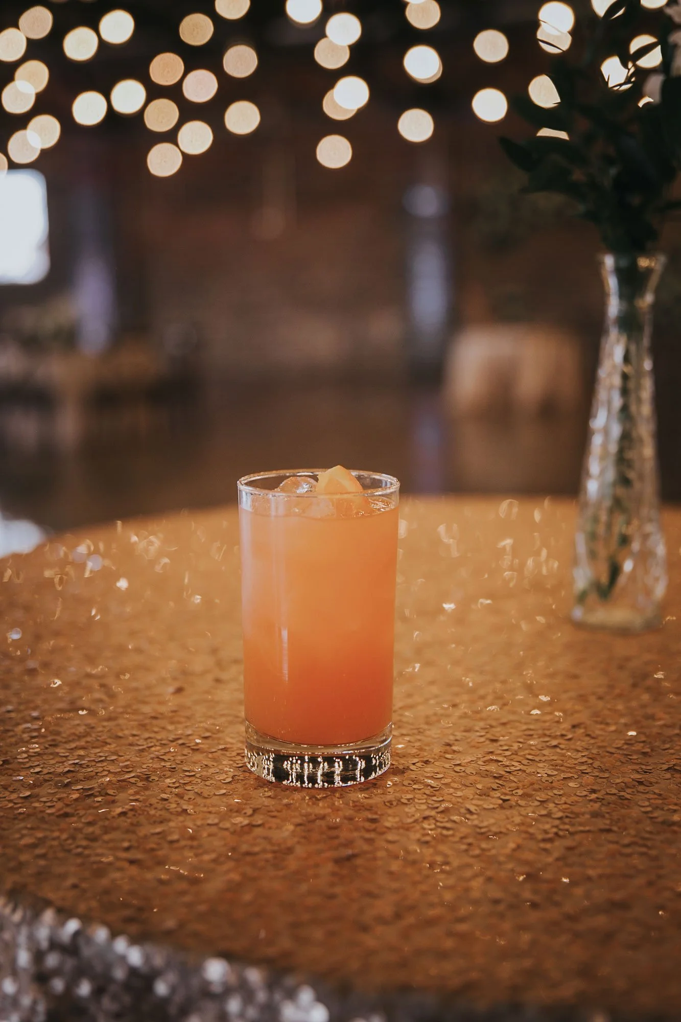 Pink drink with ice in a clear glass on a wet, reflective table. In the background, blurred lights and a vase with dark flowers.