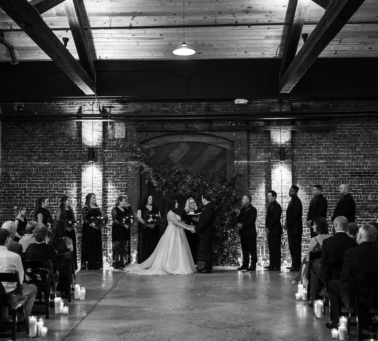 Black and white photo of a wedding ceremony with a bride and groom exchanging vows in front of officiant, with bridesmaids and groomsmen standing on either side, in a venue with brick walls and wooden ceiling beams.