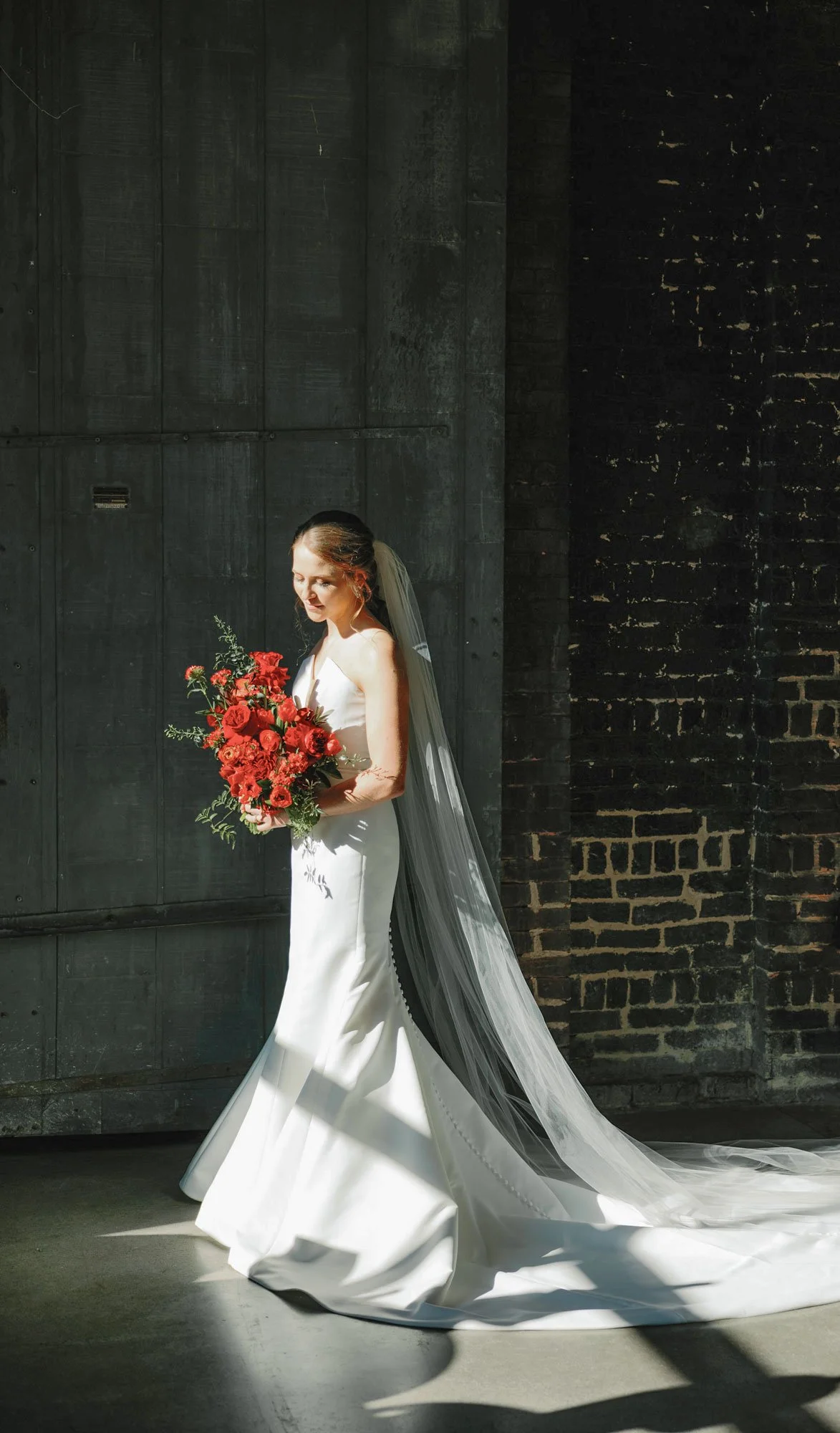 Bride in white wedding gown holding a bouquet of red flowers, standing indoors with dark gray wall and brick wall background, with sunlight casting shadows.