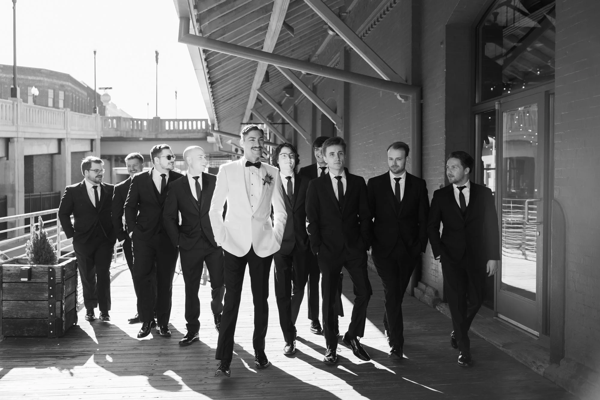 A group of men in tuxedos, including one in a white jacket, walking on a wooden deck outside a building, with some smiling and others looking serious, during the daytime.