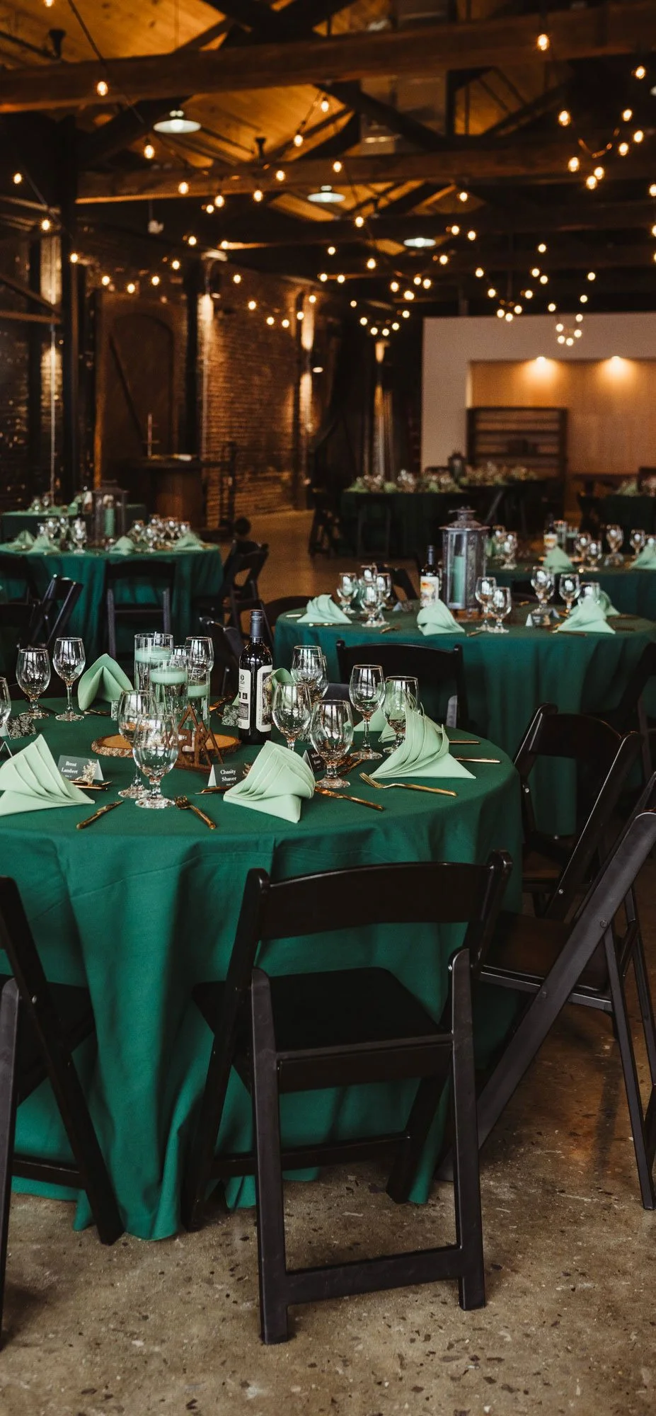 Wedding venue setup with round tables draped in green tablecloths, arranged with wine glasses, napkins, and gold flatware, set inside a rustic brick venue with string lights hanging from the ceiling.