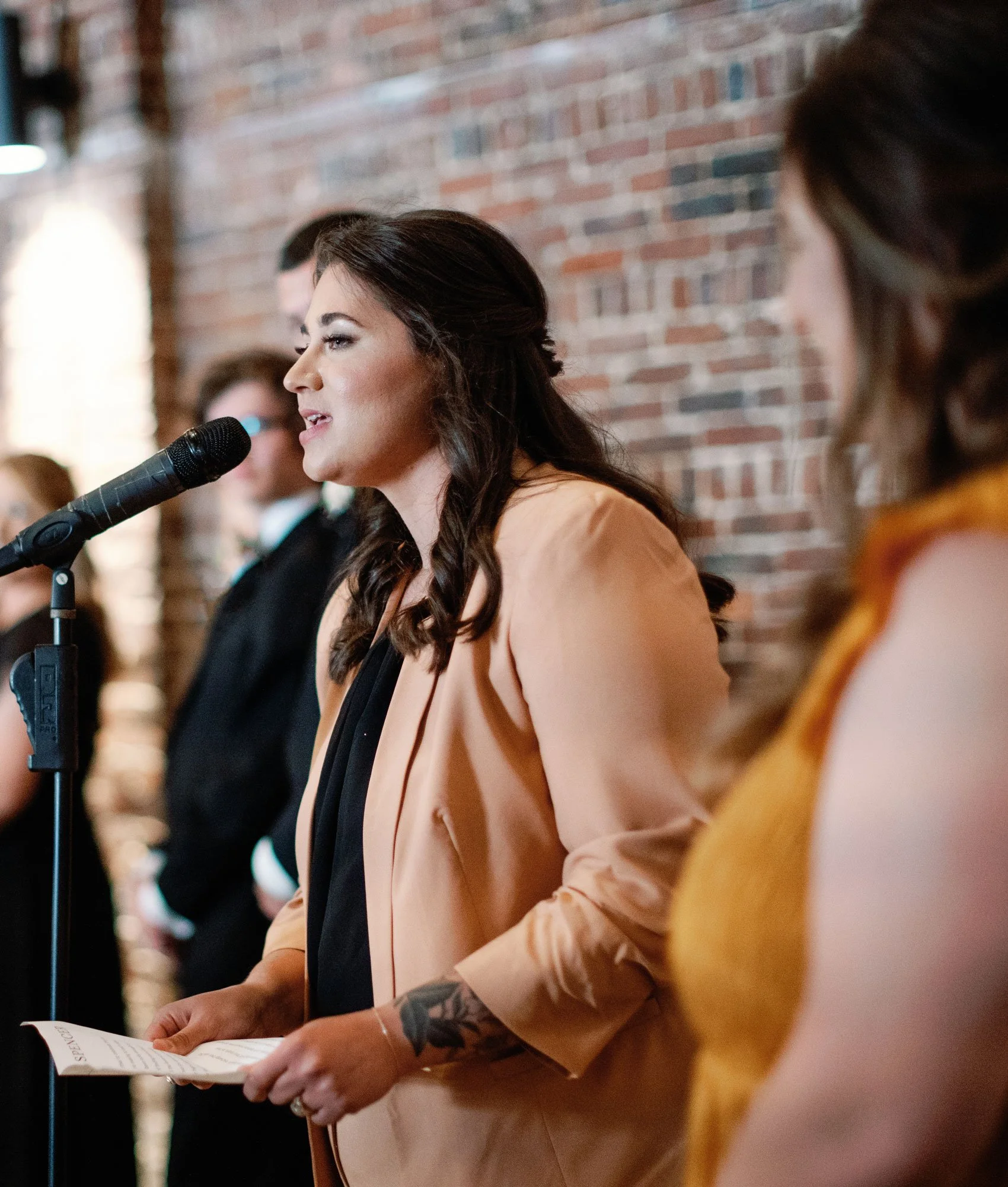 A woman with dark, wavy hair speaking into a microphone at a formal event, holding a paper, with a brick wall in the background and others standing nearby.