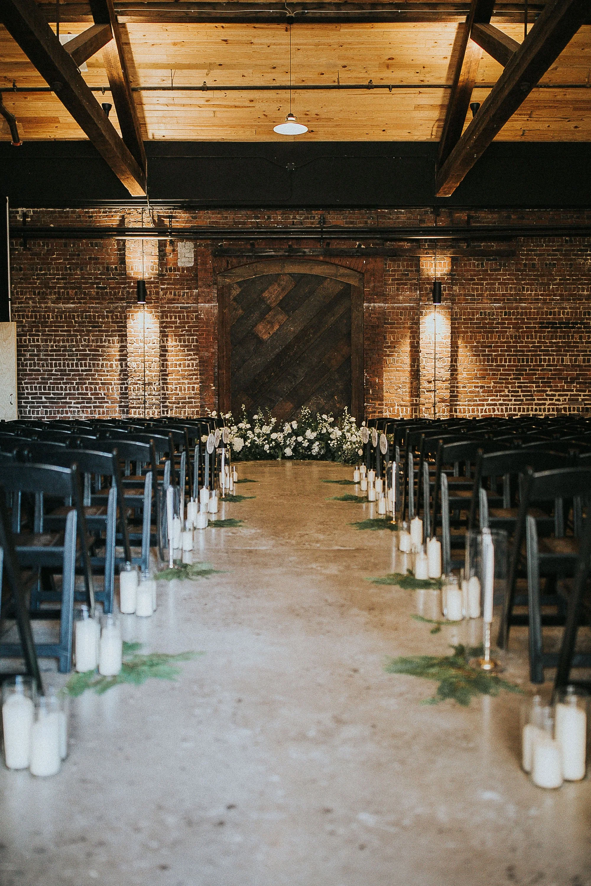 An indoor Downtown Knoxville wedding ceremony setup with rows of black chairs on either side of a central aisle, decorated with white candles and greenery, leading to a floral arrangement and a large rustic wooden door on a brick wall.