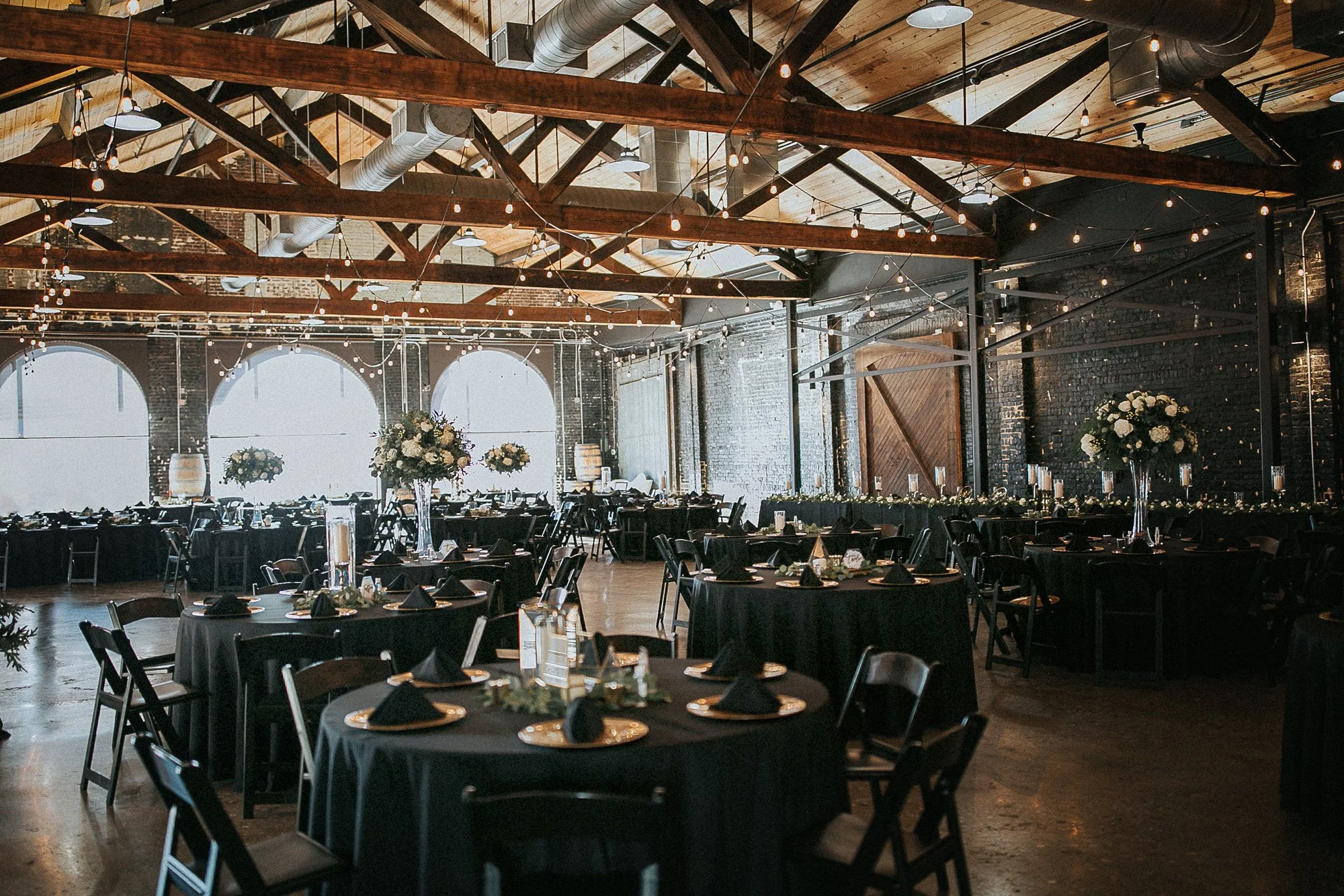 Indoor Knoxville Venue with round tables covered in black tablecloths, black napkins, and gold chargers, decorated with floral centerpieces of white flowers, with exposed brick walls, wooden ceiling beams, and string lights hanging overhead.