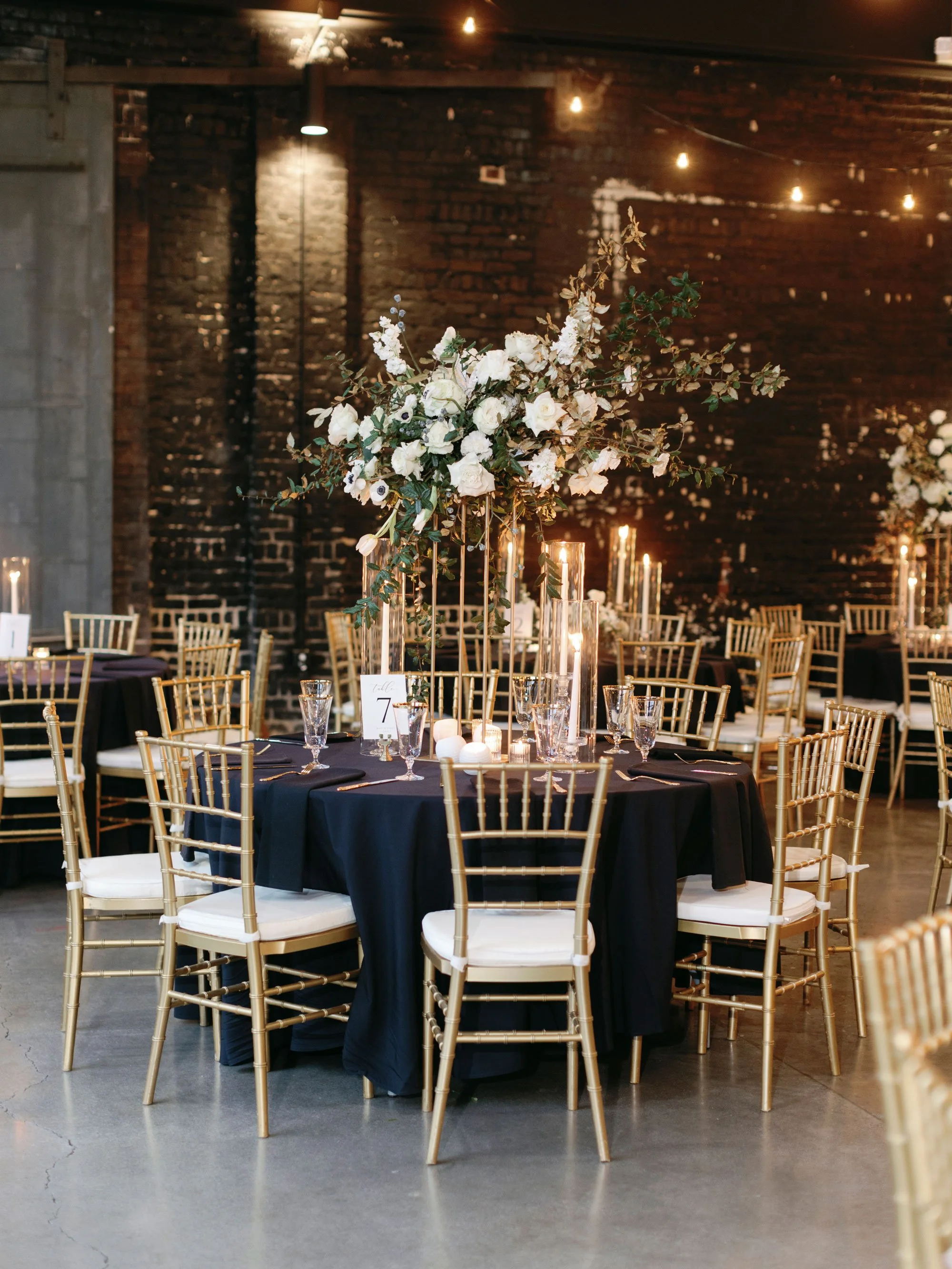 Knoxville wedding reception table with black tablecloth, gold chairs, tall floral centerpiece of white flowers and greenery, and candle decorations in a dimly lit venue with exposed brick wall.