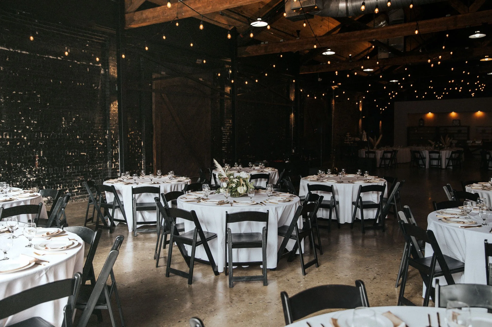 Event space with round tables covered in white tablecloths, each set with glassware, plates, and napkins, and decorated with floral centerpieces, inside a rustic venue with exposed brick walls, wooden beams, and string lights overhead.