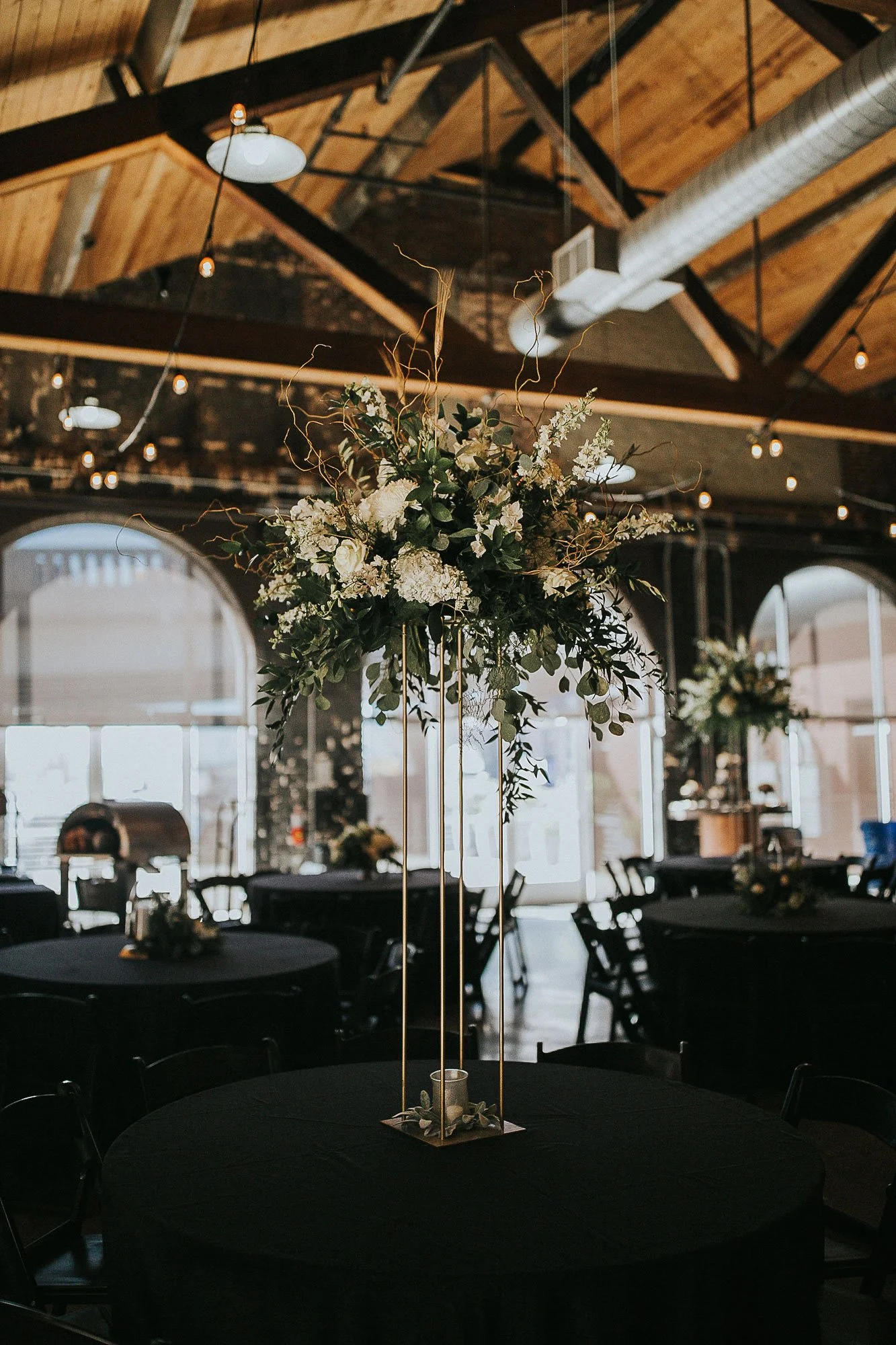Table centerpiece for a Knoxville Event Space with a tall floral arrangement of white flowers and greenery in a gold stand at a reception or event venue.