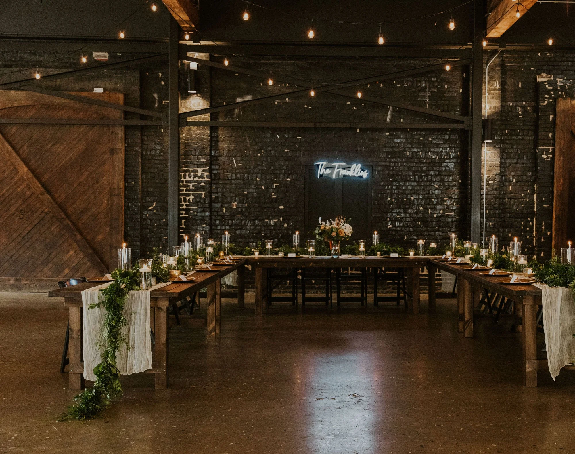 A long wooden dining table at a Downtown Knoxville event space decorated with greenery, candles, and place settings in a rustic venue with exposed brick walls and string lights