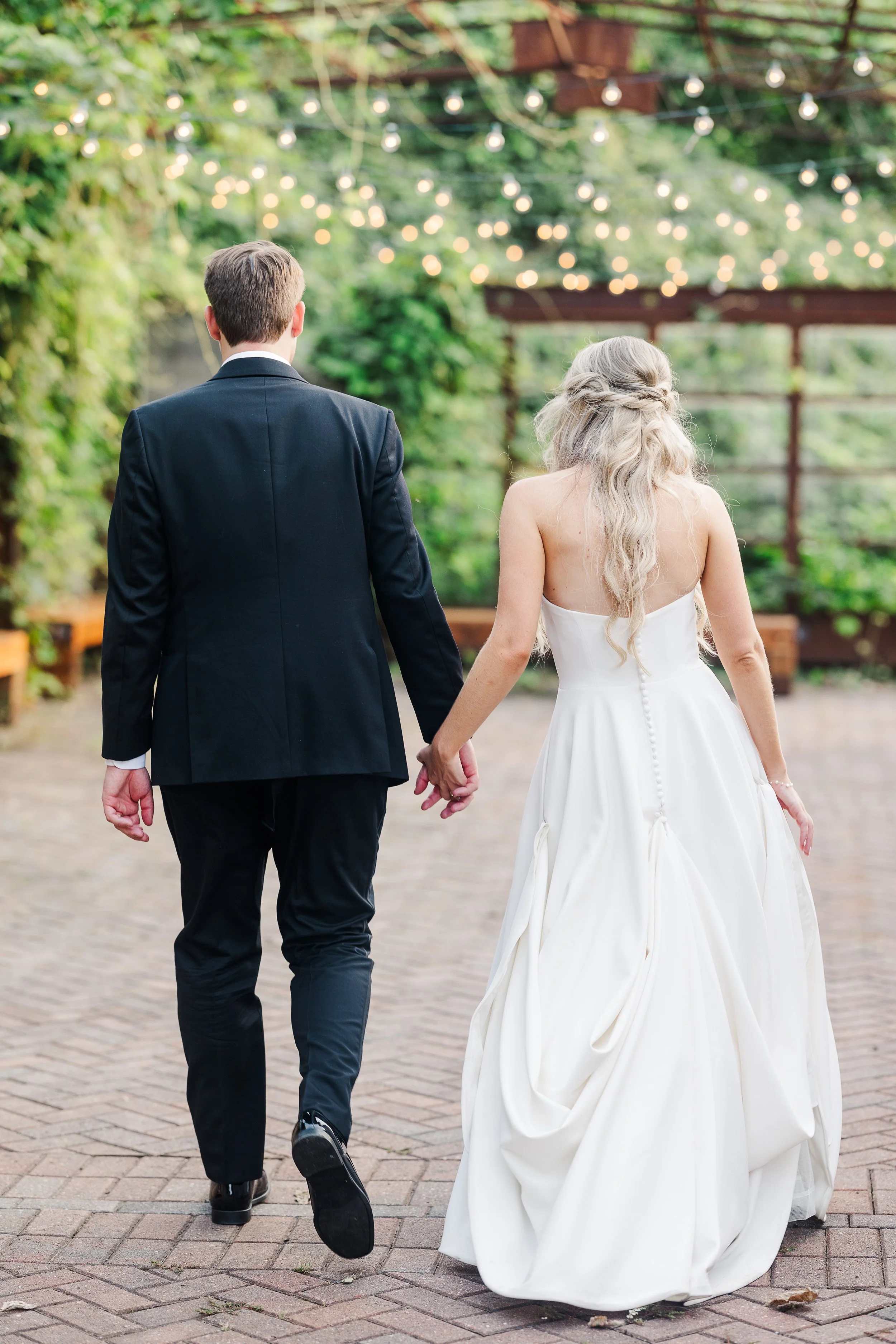 A couple dressed in wedding attire, holding hands and walking away on a paved pathway at an wedding venue in The Old City Knoxville