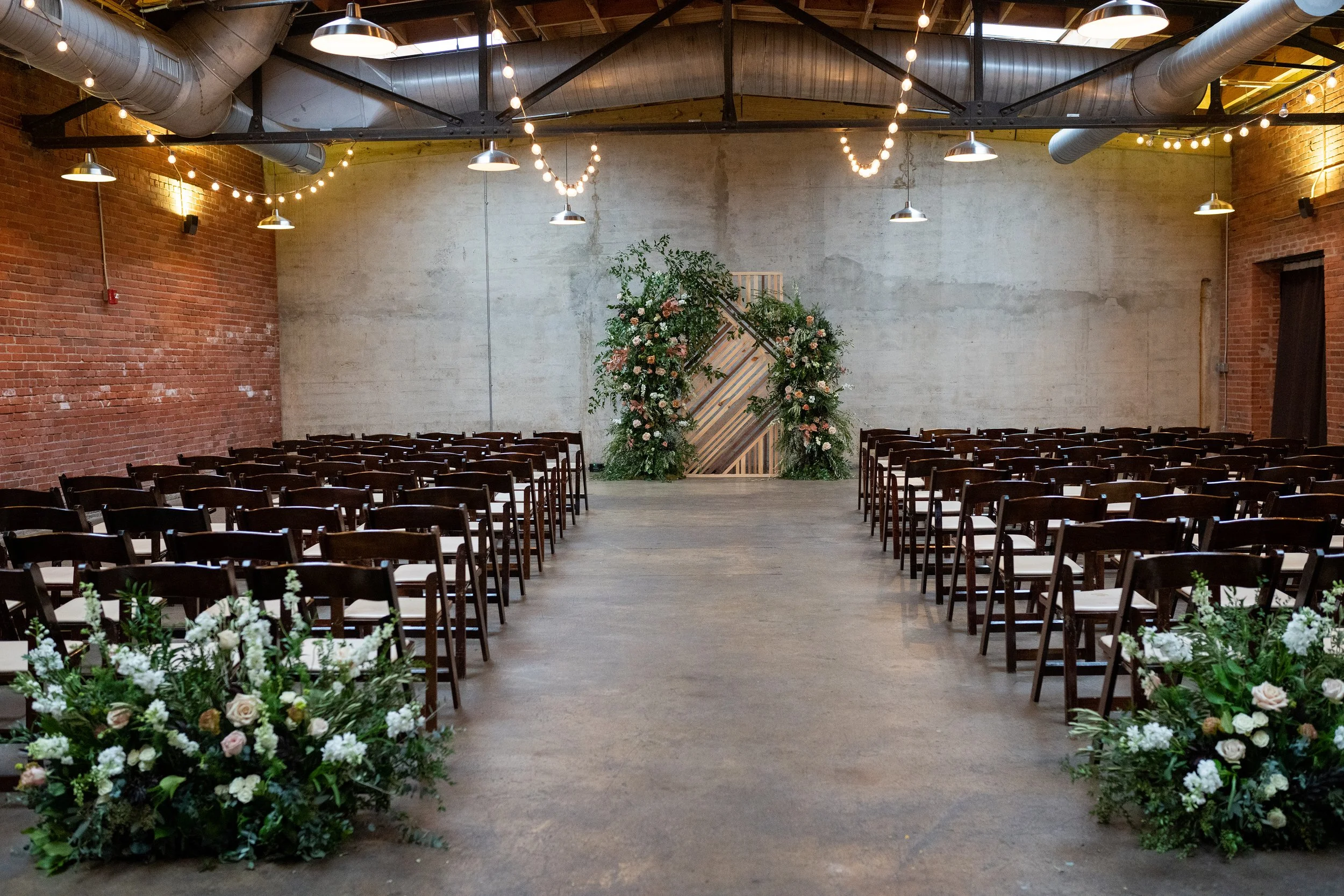 Indoor wedding ceremony venue decorated with floral arrangements and a floral arch at the front, with rows of chairs on either side. String lights hang from the ceiling over the space, which features exposed brick and concrete walls.