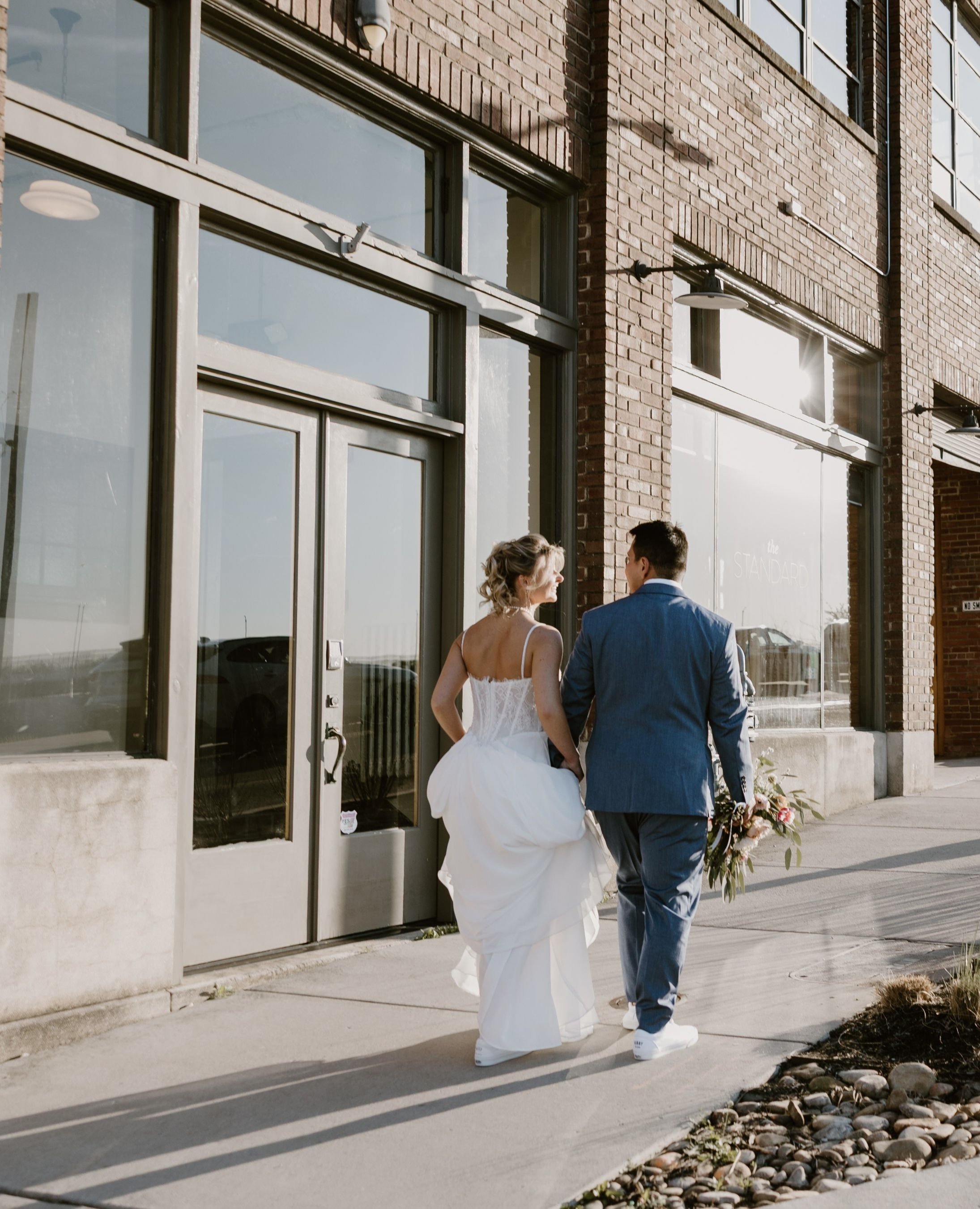 Couple dressed in wedding attire walking outside a brick building in Downtown Knoxville with large windows, sunlight reflecting off the glass.