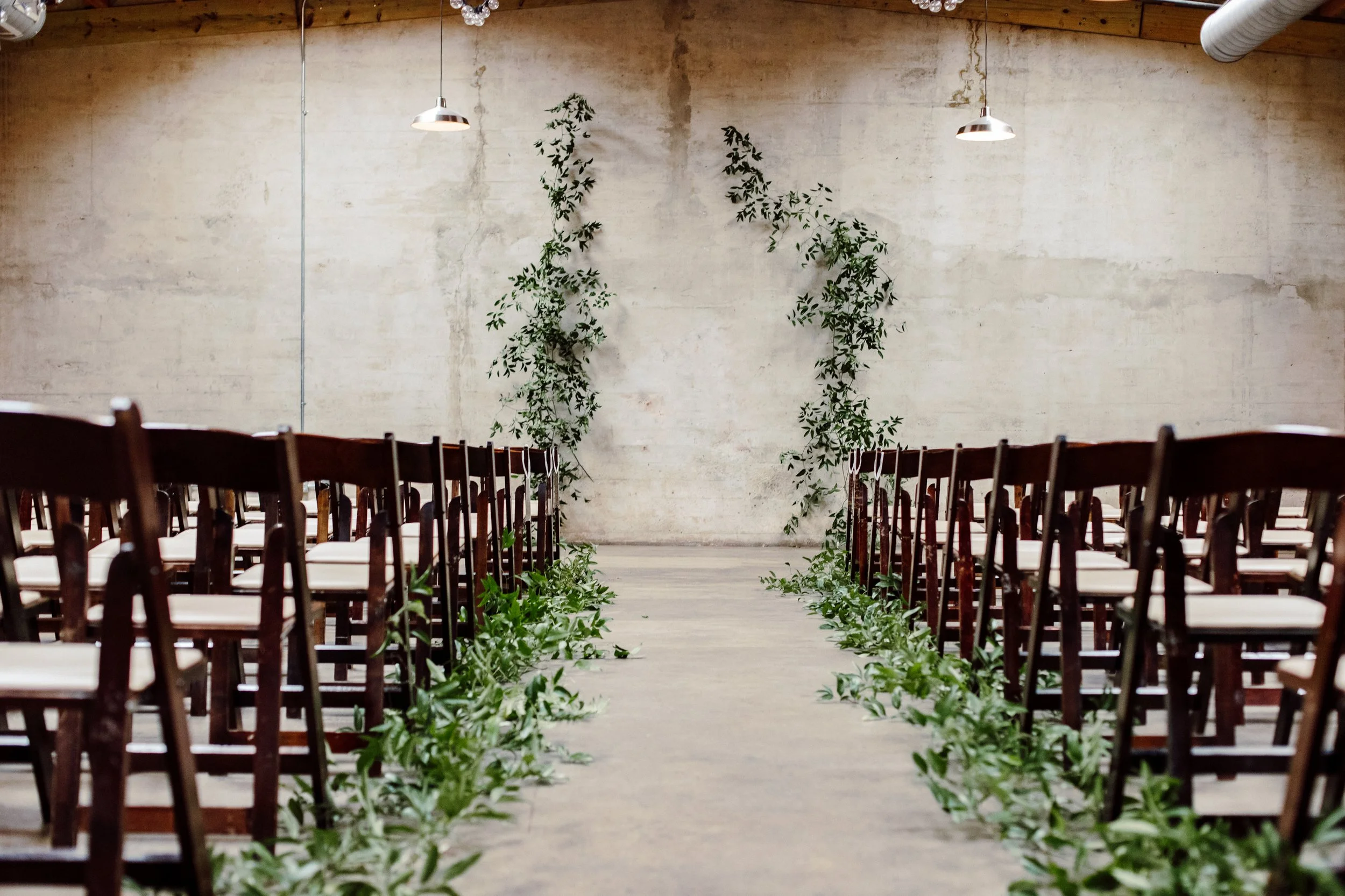 Indoor space arranged for a ceremony with rows of wooden chairs decorated with leafy greenery along the aisle. Two wall-mounted lamps and some greenery vine decor hang against a plain, unfinished concrete wall.
