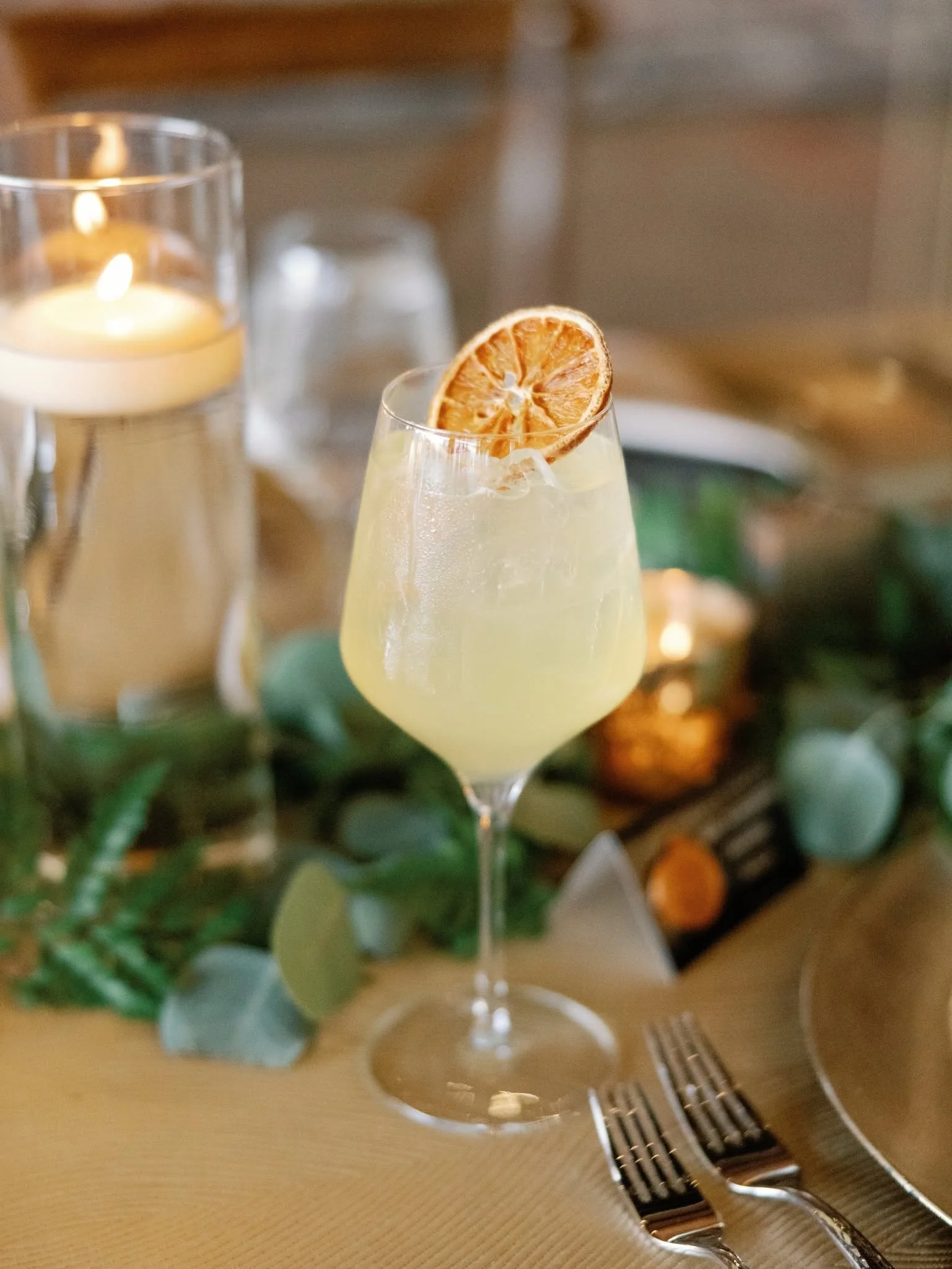 A glass of pale yellow cocktail with a slice of dried orange as garnish, placed on a table with two forks nearby, with a candle and some greenery in the background.