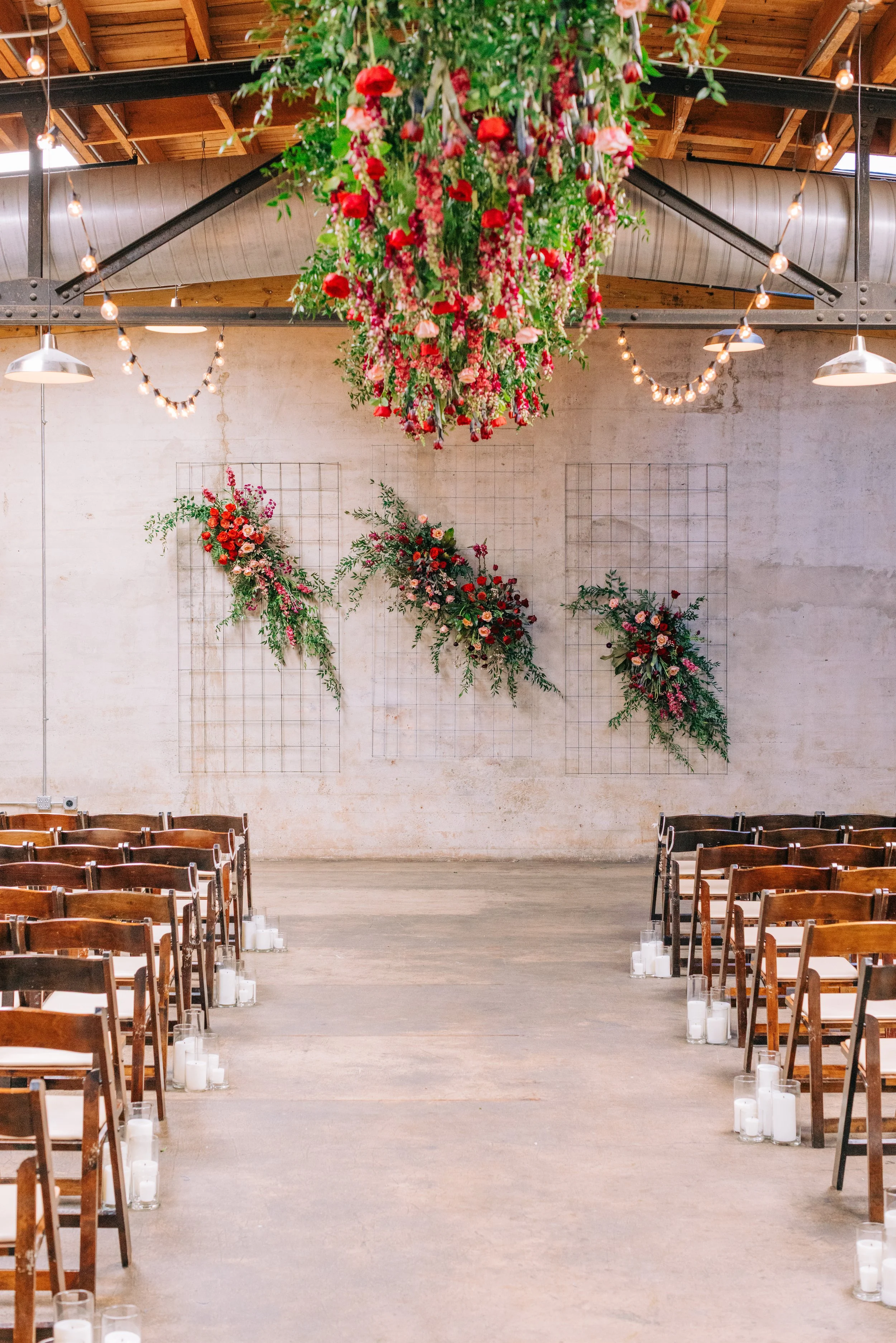 Indoor wedding ceremony setup with rows of chairs and white candles lining the aisle, a large floral arrangement hanging from the ceiling, and additional floral decorations on the back wall, in a rustic venue with string lights and wooden beams.