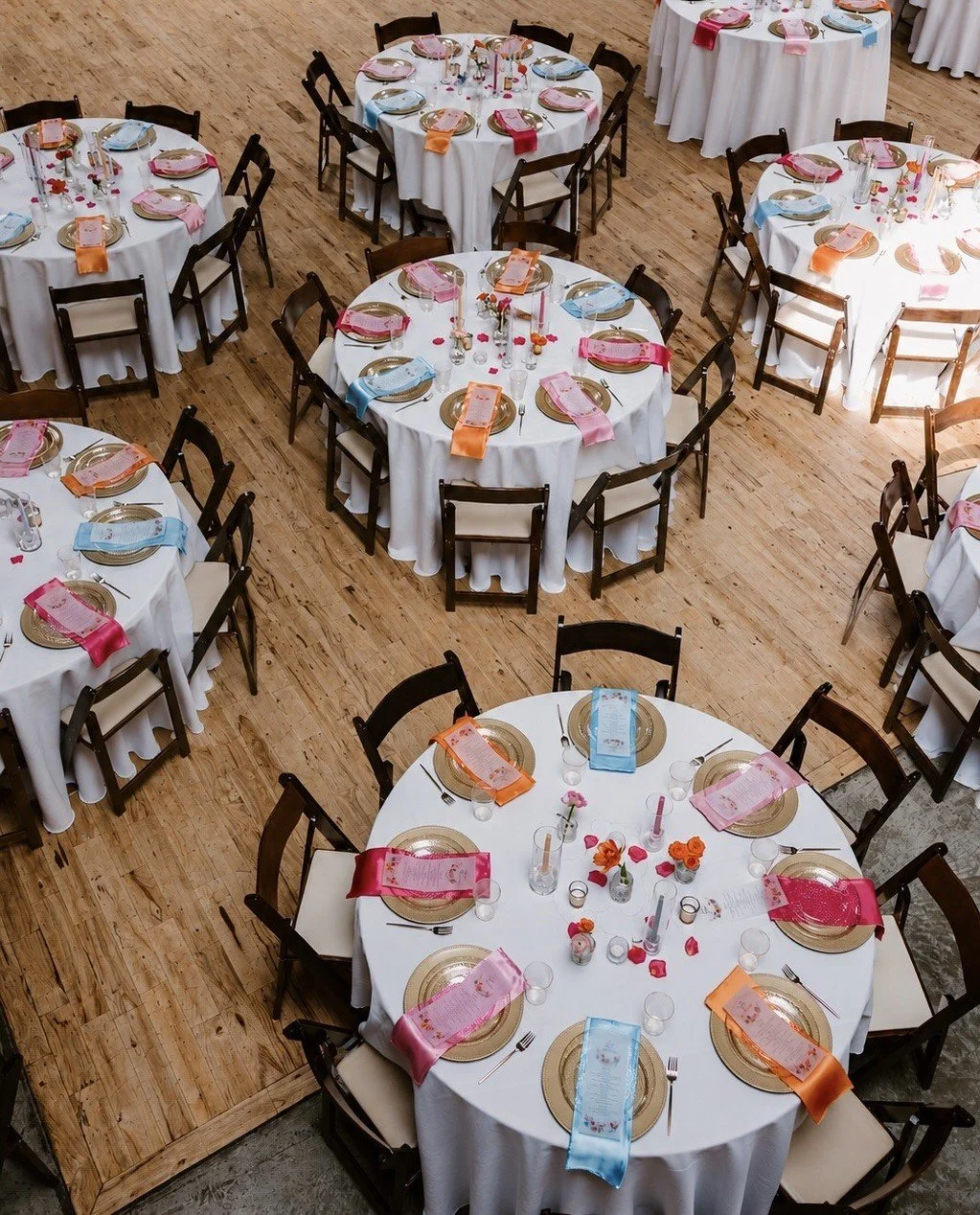 Round banquet tables with white tablecloths, set with gold chargers, pink and blue napkins, small flower arrangements, and candles, arranged in a large event hall with wooden floors.