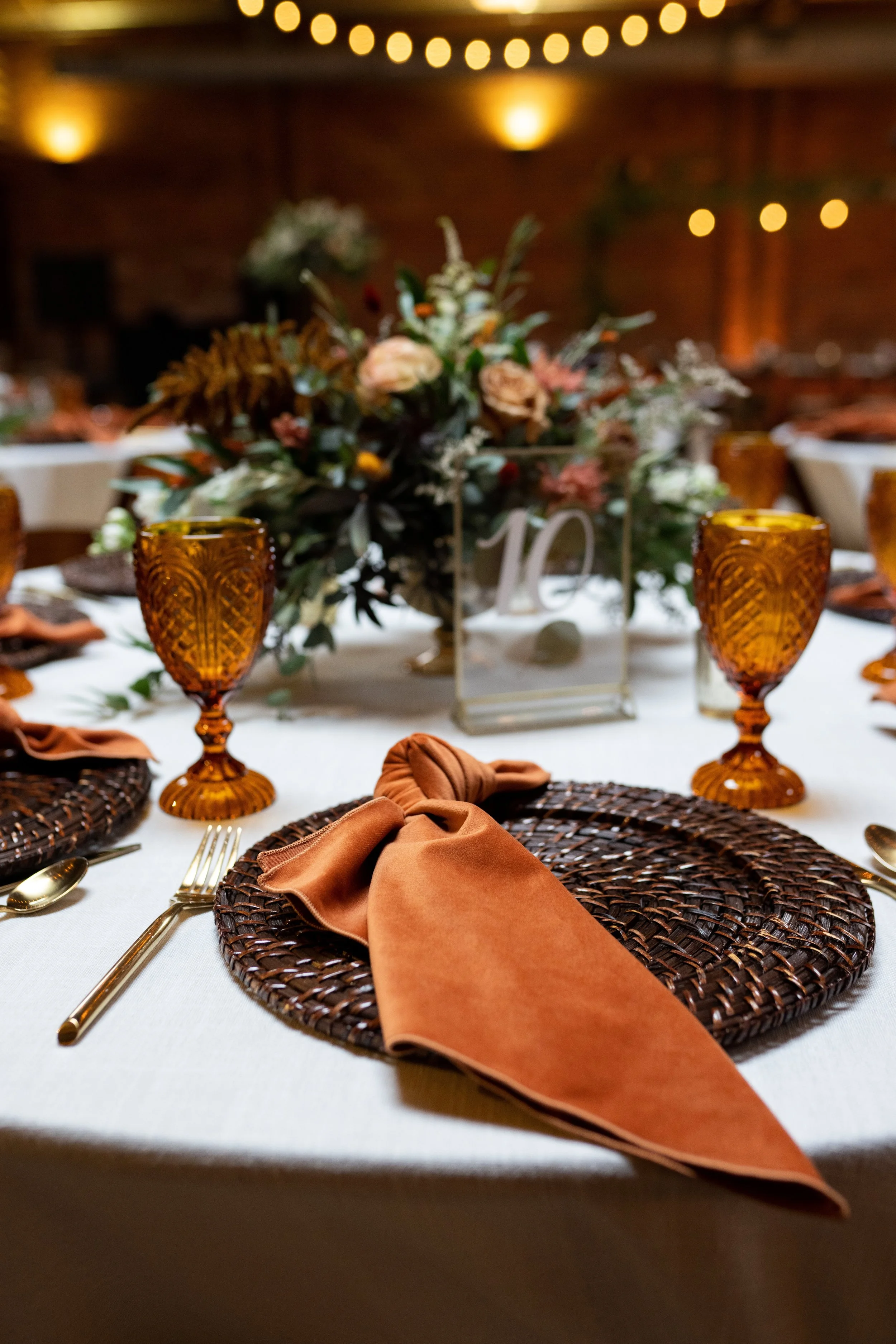 A close-up of a table setting at an elegant event, with woven brown placemats, orange napkins, gold utensils, and amber glassware, with a floral centerpiece in the background.