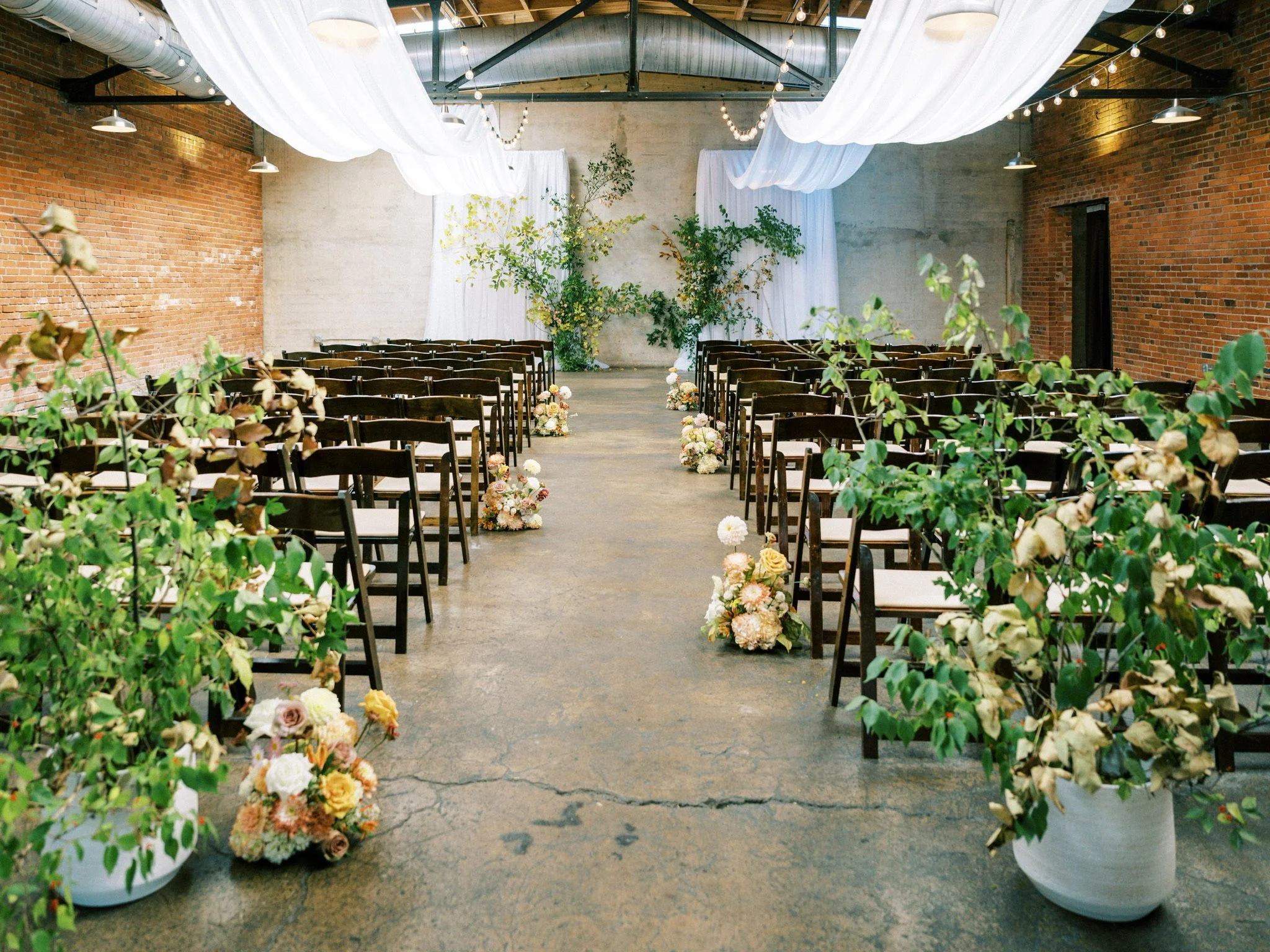 Elegant Indoor wedding ceremony setup at The Standard in Downtown Knoxville with rows of wooden chairs on either side of a decorated aisle, flanked by large white pots filled with greenery and flowers, with a backdrop of white draping.