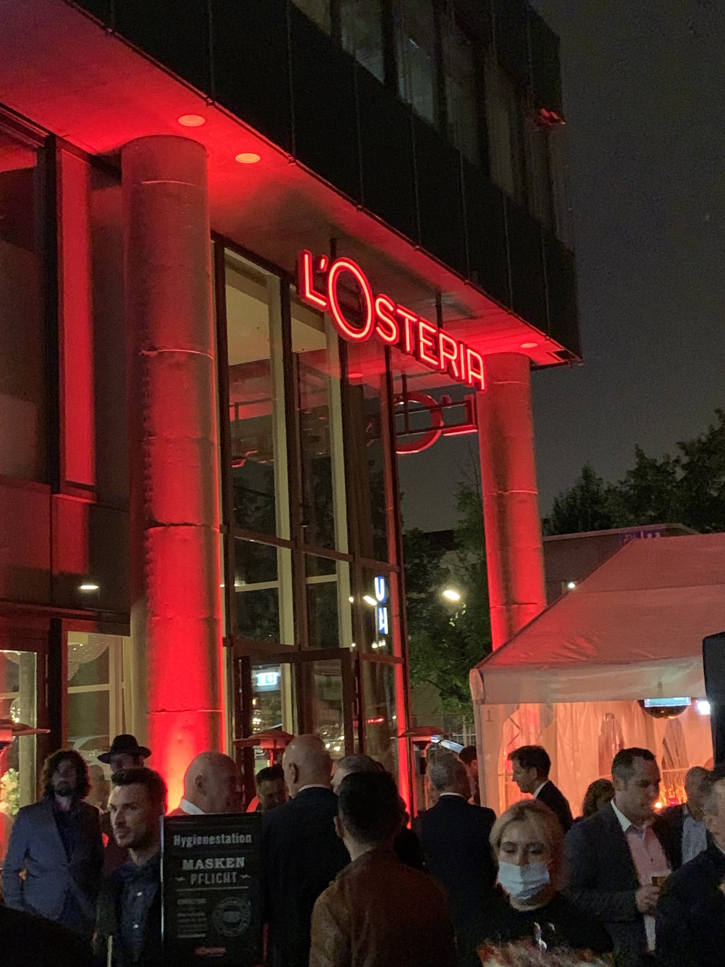 Crowd of people gathered outside a building at night, with a red neon sign reading 'L'Osteria', and some individuals wearing masks.