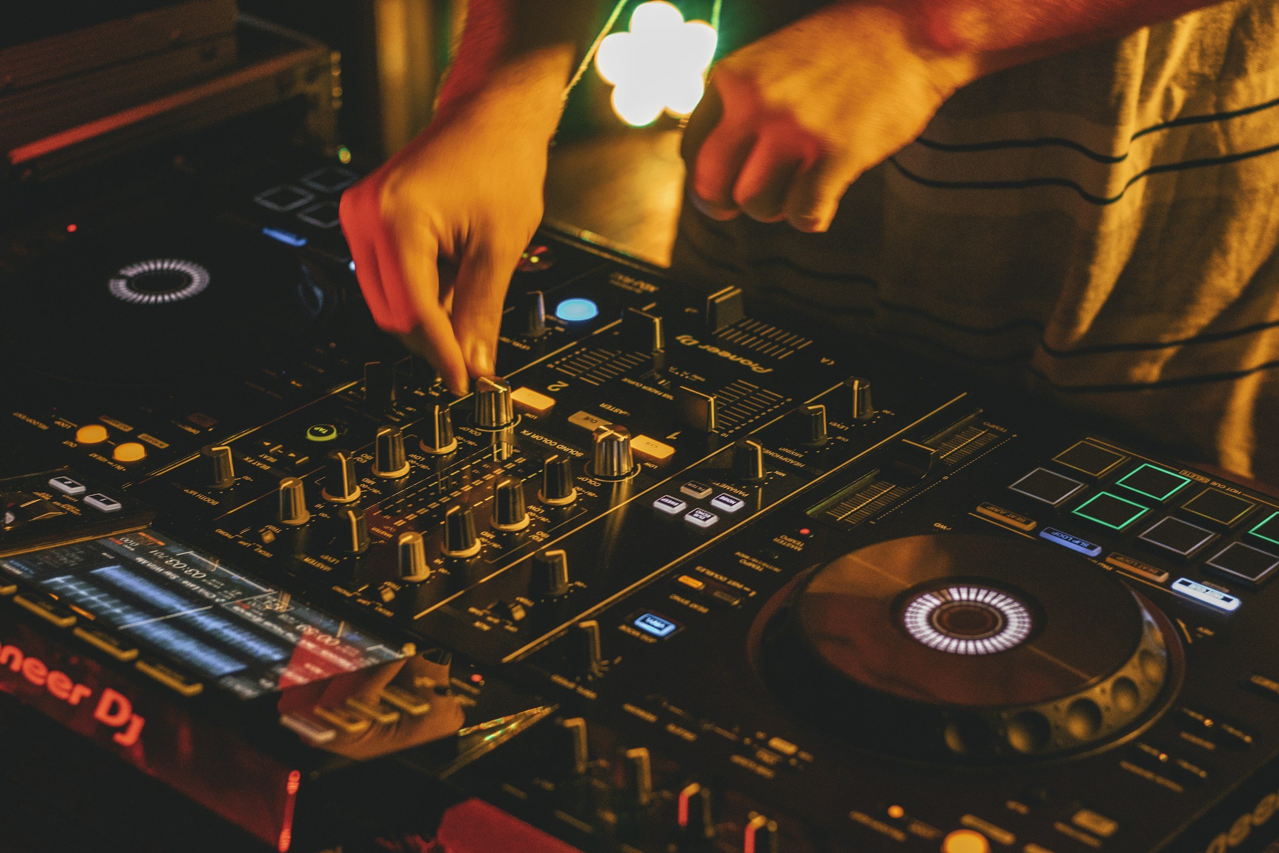 Close-up of hands operating a DJ mixing console at a party, with colorful lights illuminating the scene.