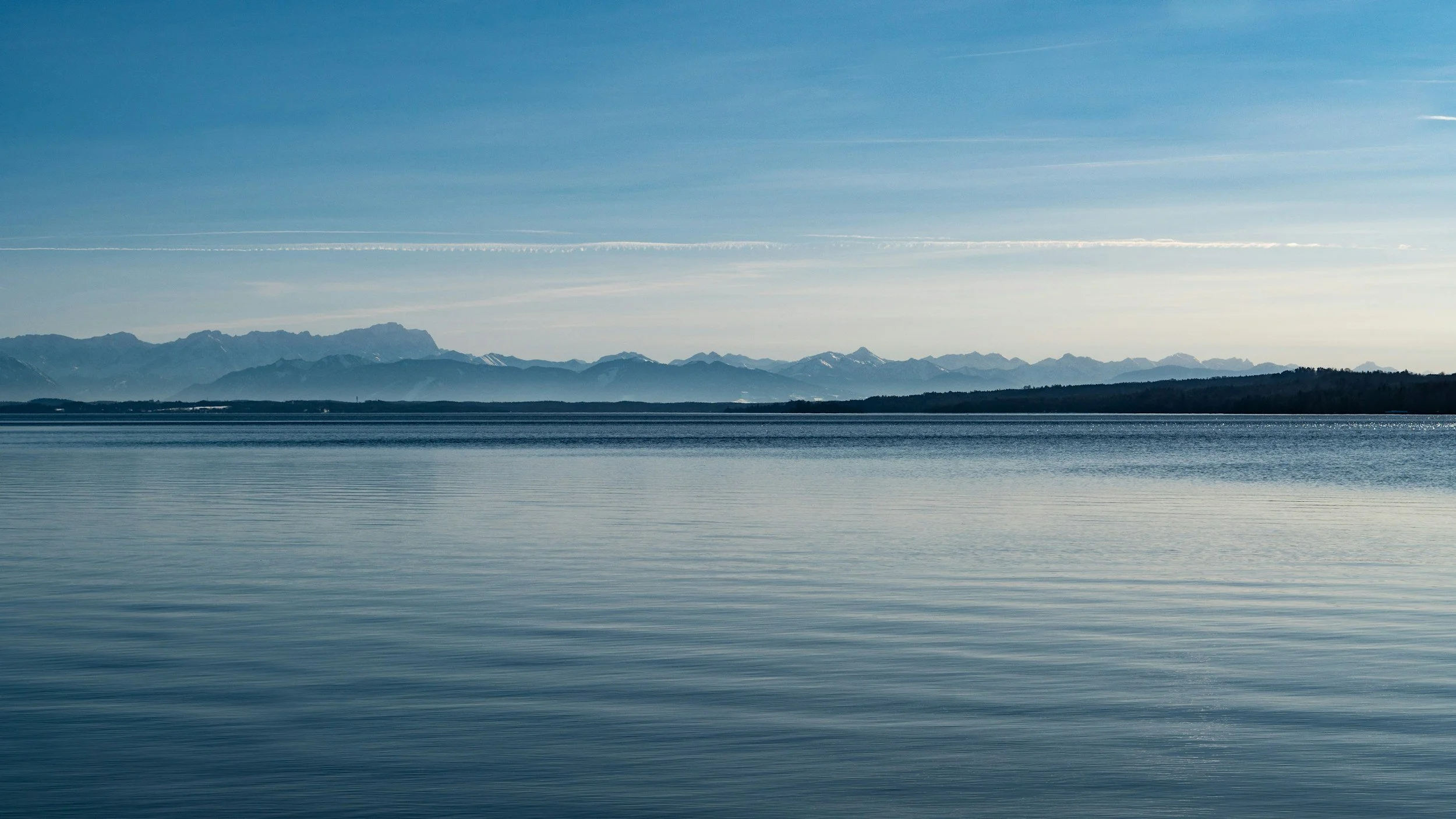 A serene body of water with distant mountain ranges under a clear blue sky, with some faint clouds and contrails.
