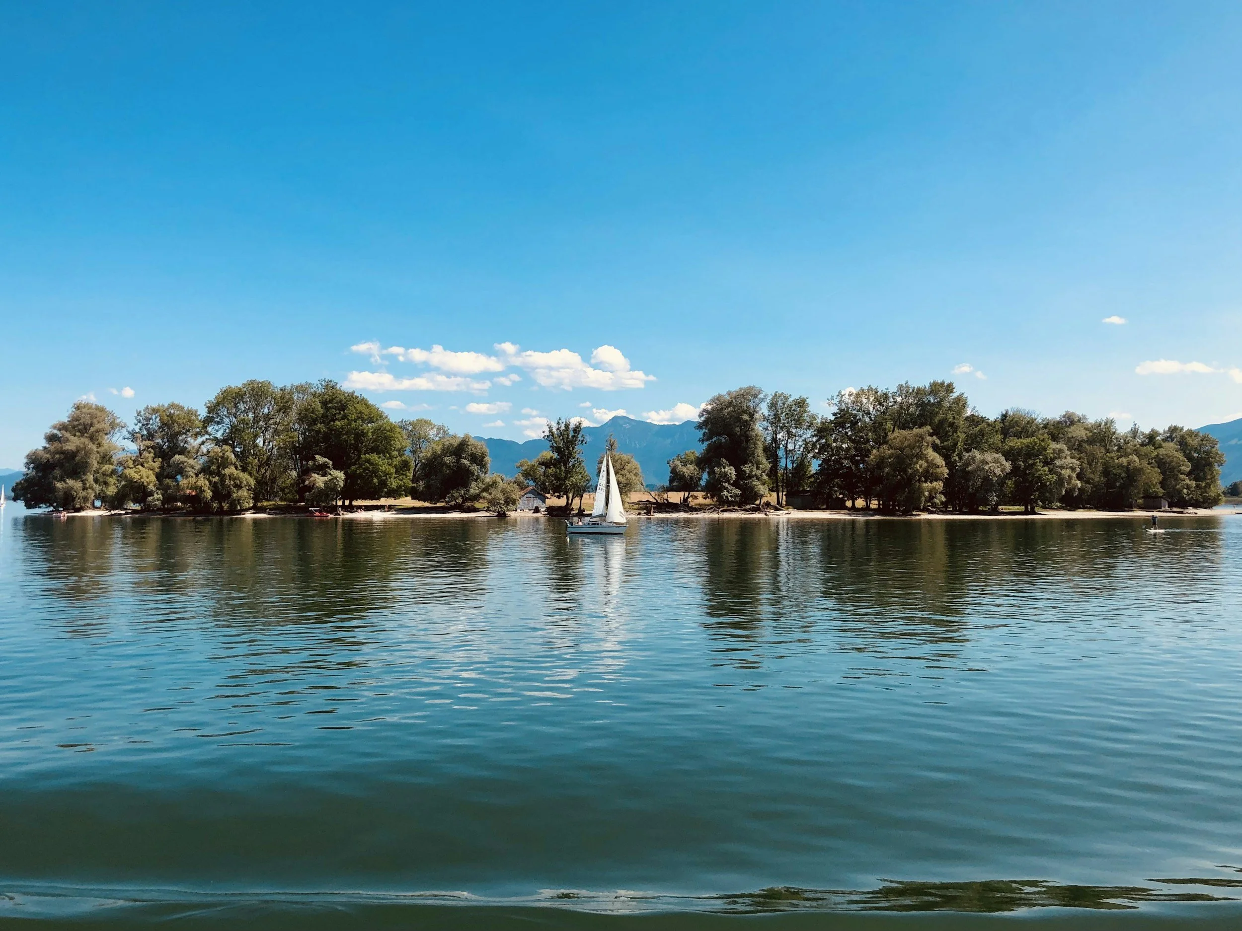 A scenic view of a lake with a sailboat sailing across calm water, surrounded by lush trees and mountains in the background under a bright blue sky with scattered clouds.