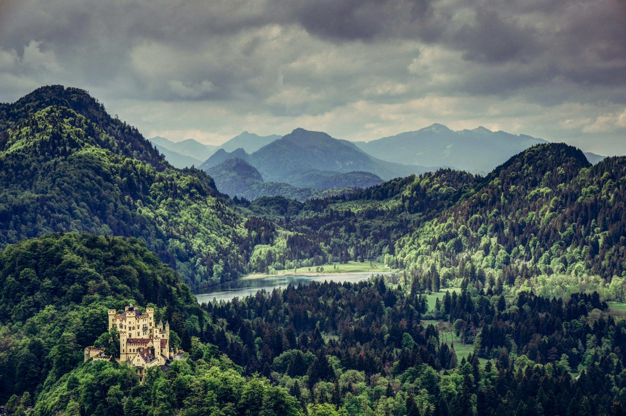 A lush green mountain landscape with a river flowing through the valley and a castle-like building on a hill in the foreground, under cloudy skies.