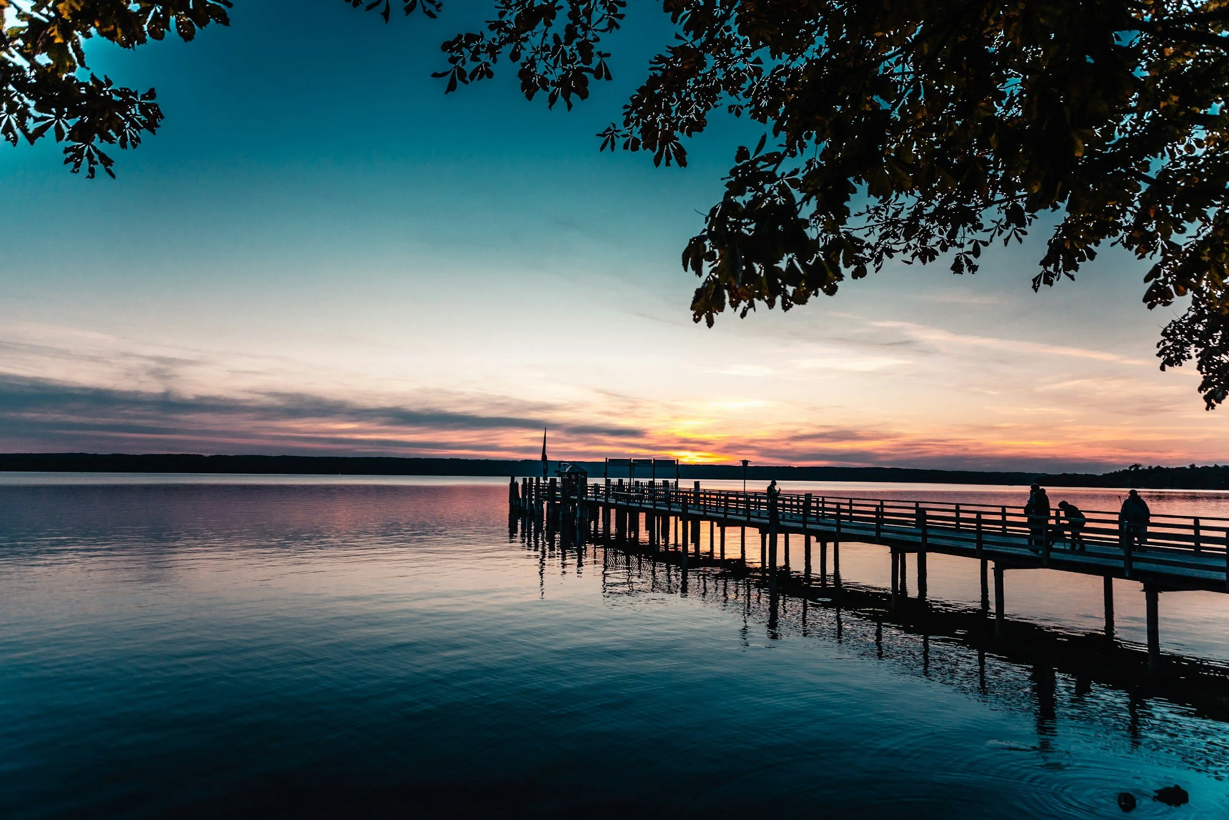 Sunset over a calm body of water viewed from a shoreline, with a pier extending into the water and trees framing the scene.