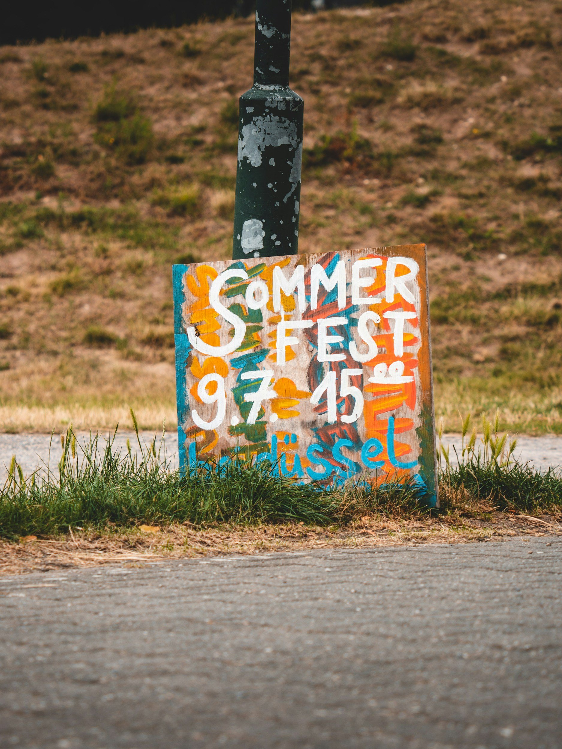 Colorful hand-painted sign with the words 'Summers Fest'. The sign is placed on the grass next to a paved road, with a hillside in the background.