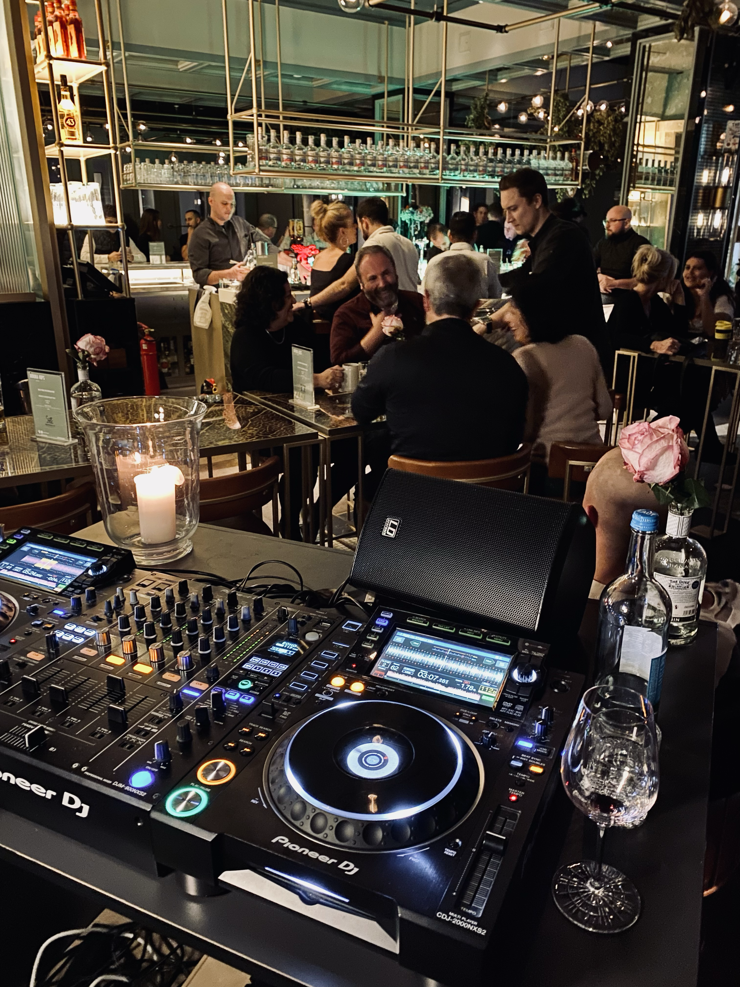 A nightclub or bar setting with people socializing around a bar and seated at tables, a DJ equipment setup with mixers and turntables in the foreground, and glasses and bottles on the tables, illuminated by warm and dim lighting.