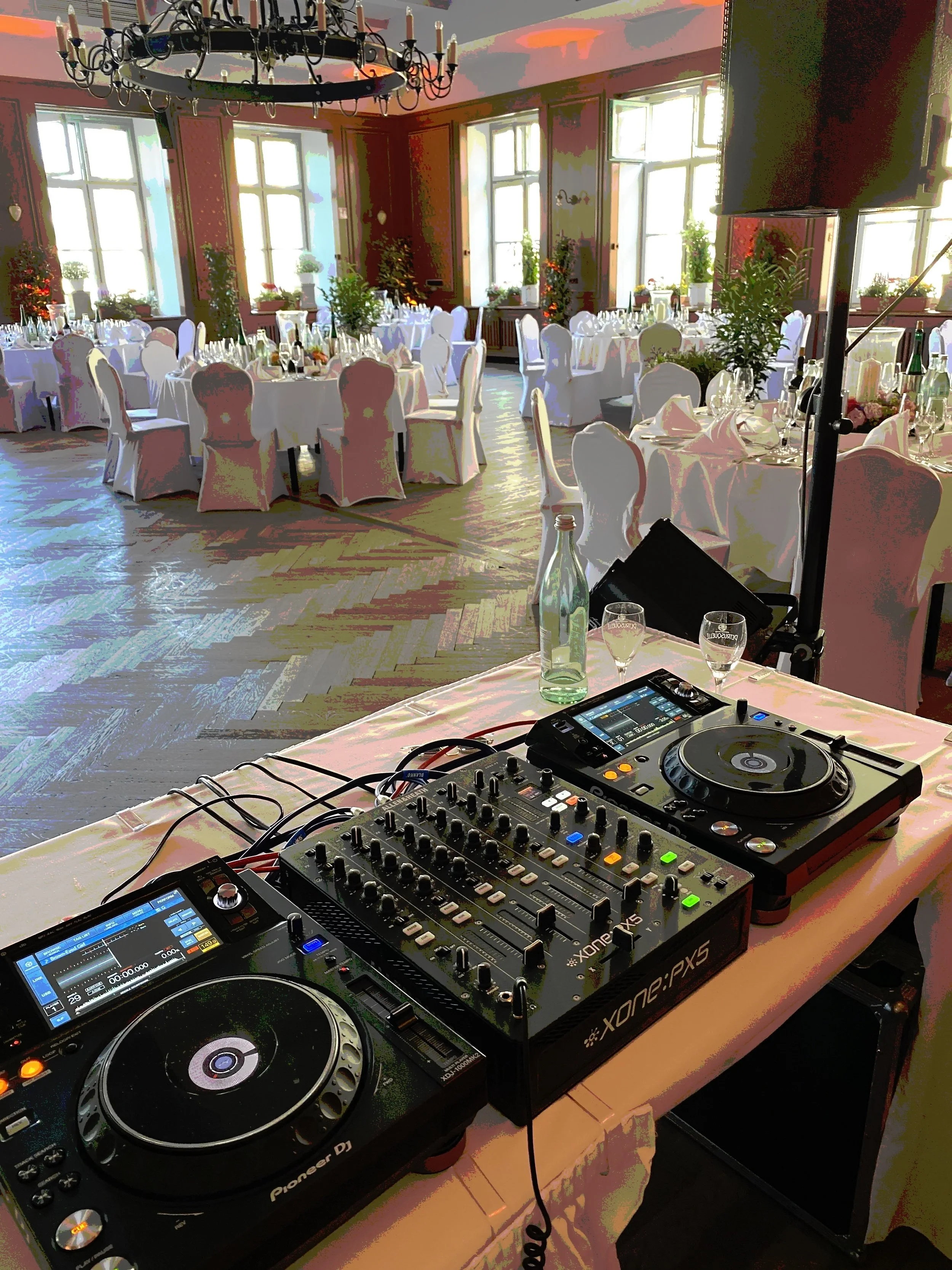 A DJ setup with two Pioneer DJ controllers, a mixer, and a touchscreen device on a table, in a decorated banquet hall with round tables, white chairs, and large windows.