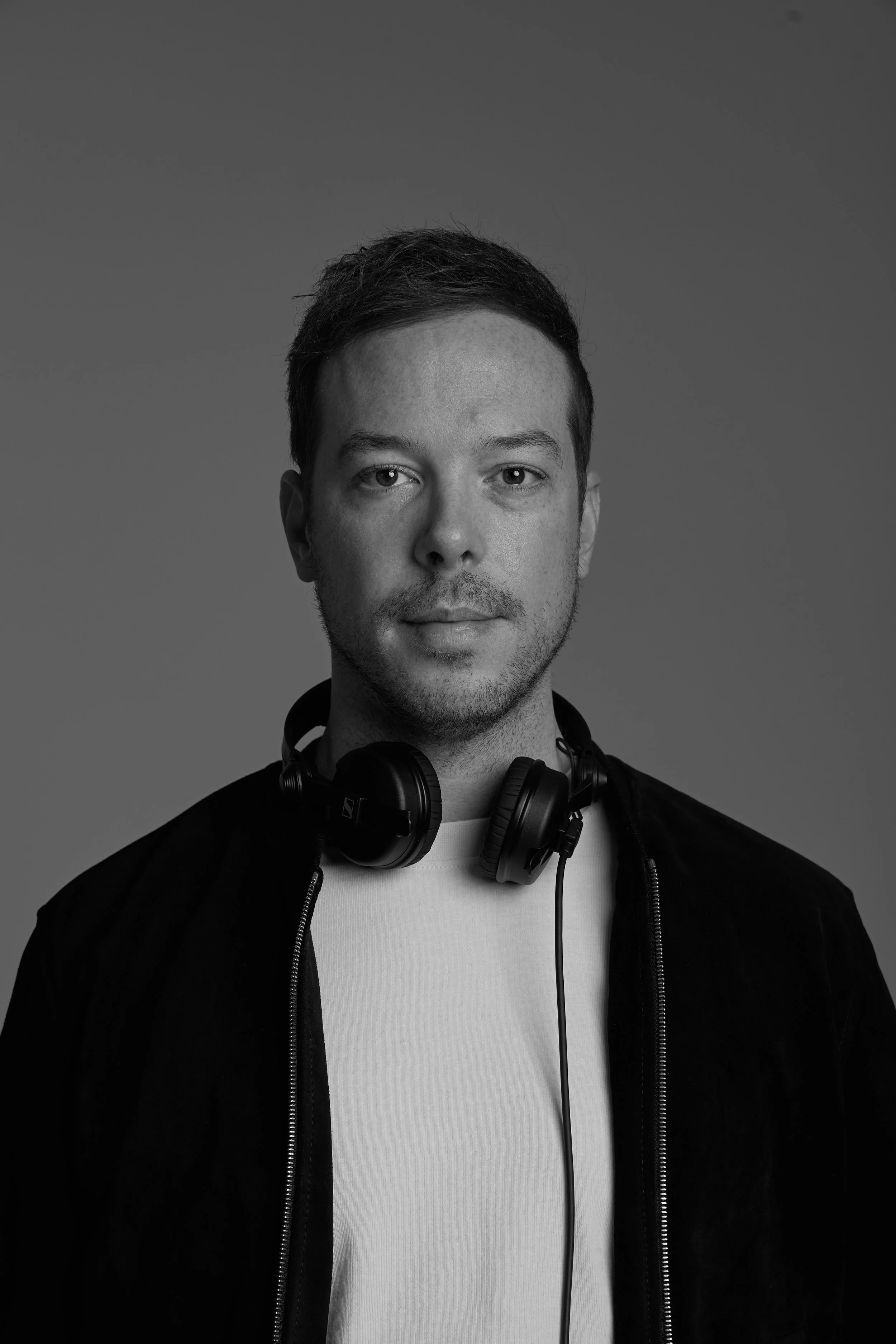 Black and white photo of a young man with short hair, wearing a white t-shirt and a black jacket with a zipper, and headphones around his neck, against a plain gray background.