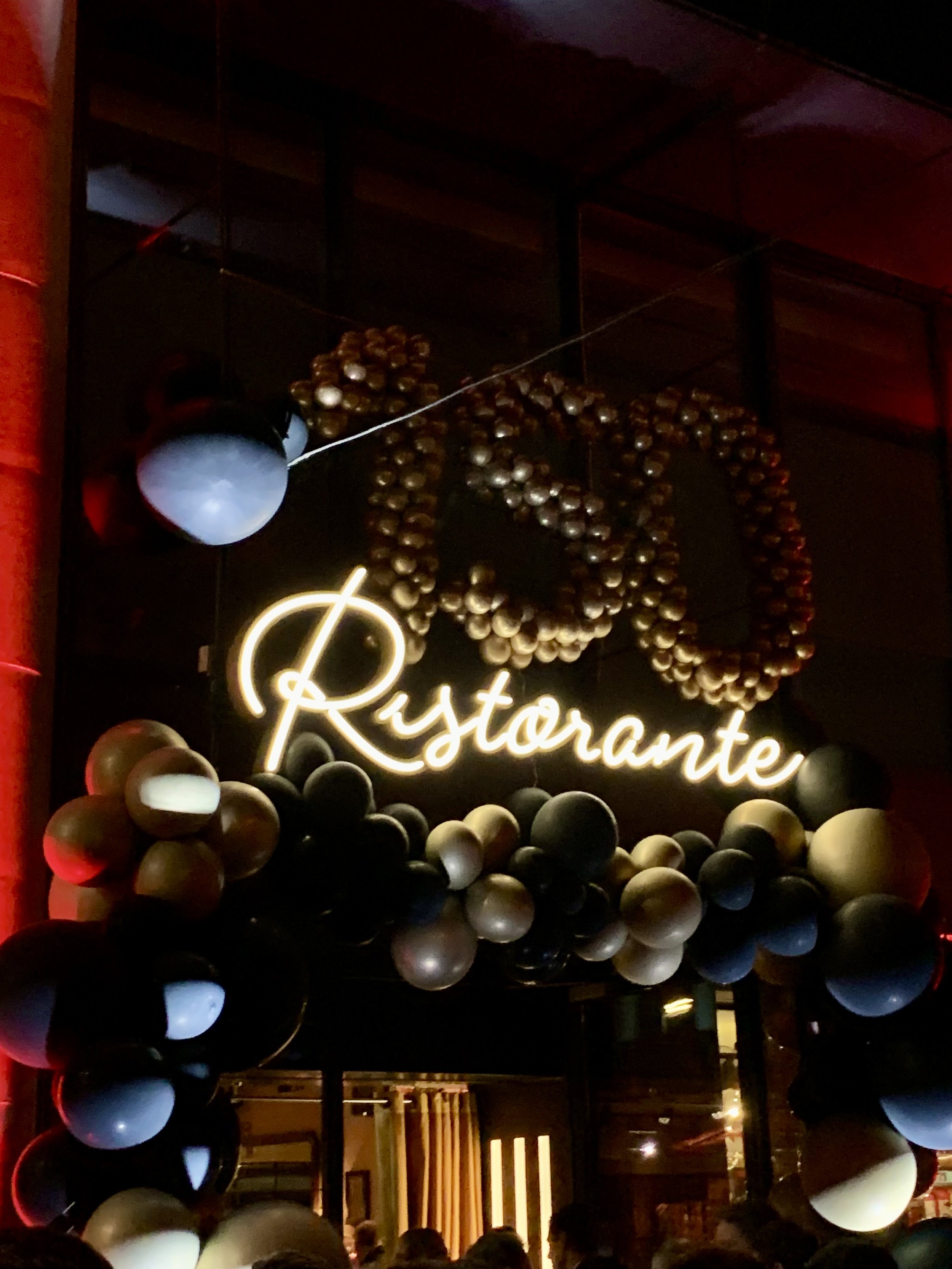 Decorative sign displaying the word 'Restaurante' with a floral wreath made of small balloons around it, and a column of inflated balloons along the sides, in a dimly lit restaurant or bar setting.