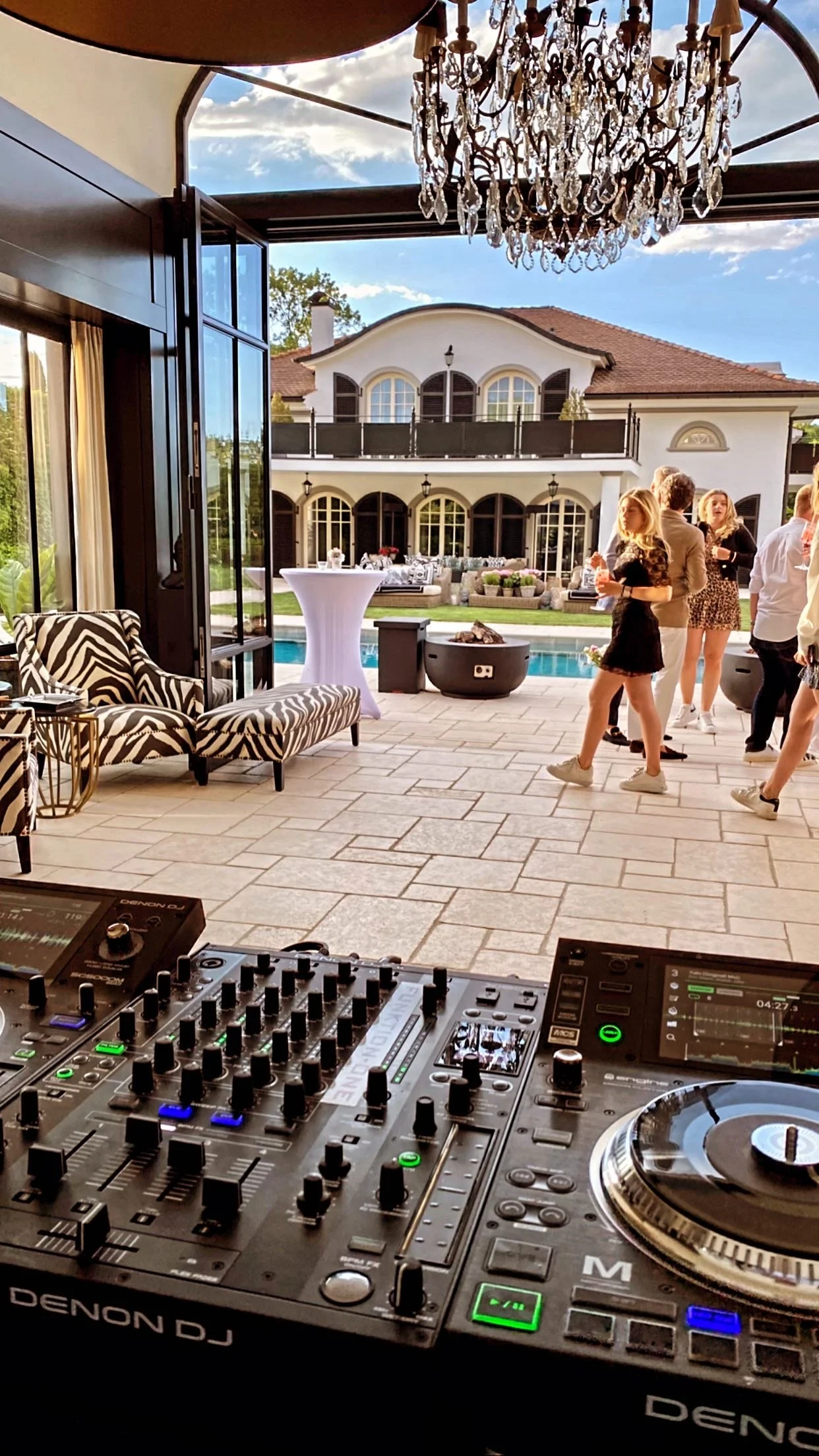 Inside a luxurious house, there is a DJ setup in the foreground with turntables and mixer. In the background, guests are socializing near a pool on a patio, with a large, elegant house featuring arched windows and a balcony, under a blue sky with clouds. A chandelier hangs from the ceiling, and zebra print lounge chairs are visible on the left.