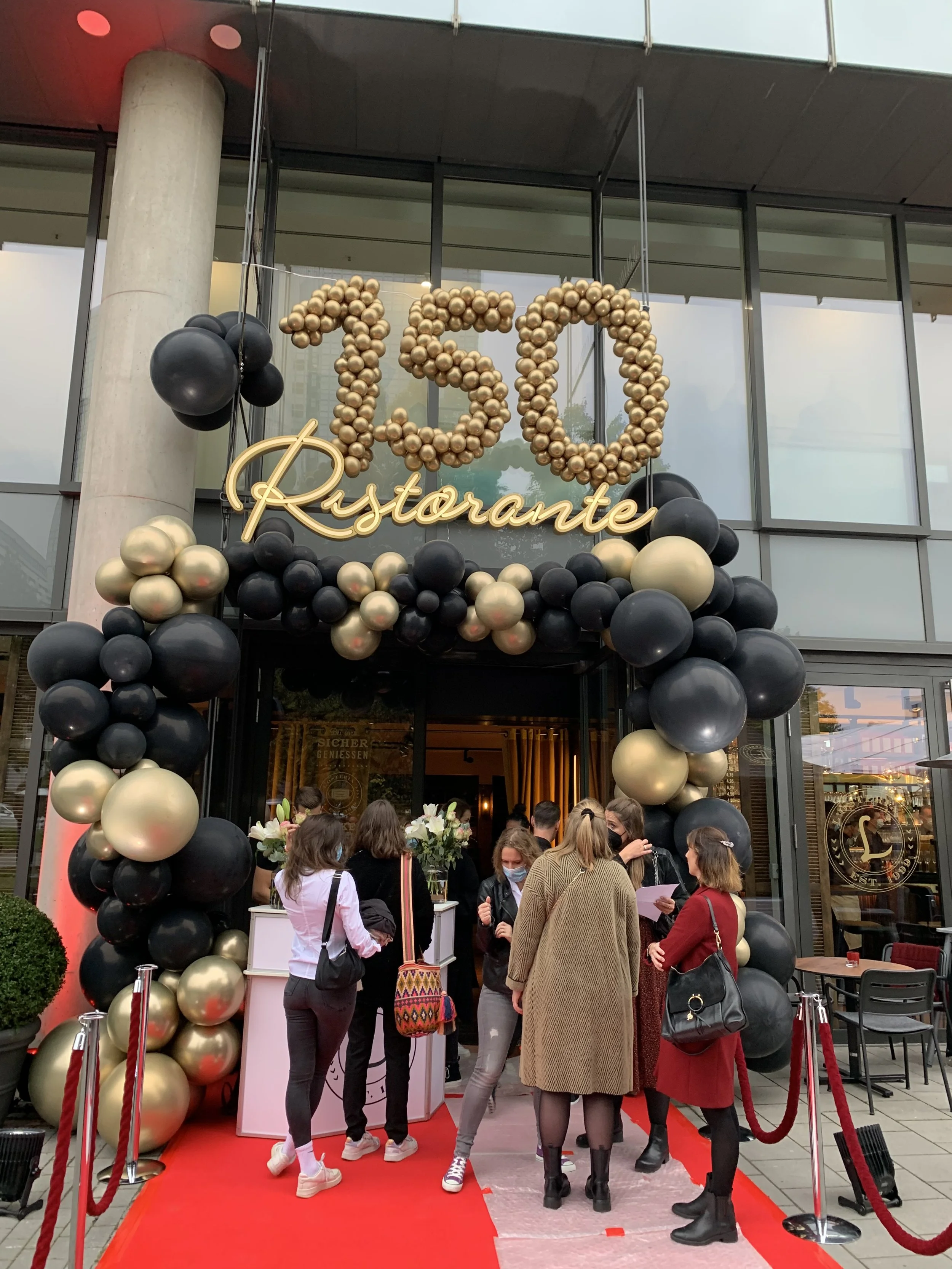 People gathered outside a restaurant entrance adorned with a balloon arch and a sign that reads '73,' with a red carpet and velvet ropes.