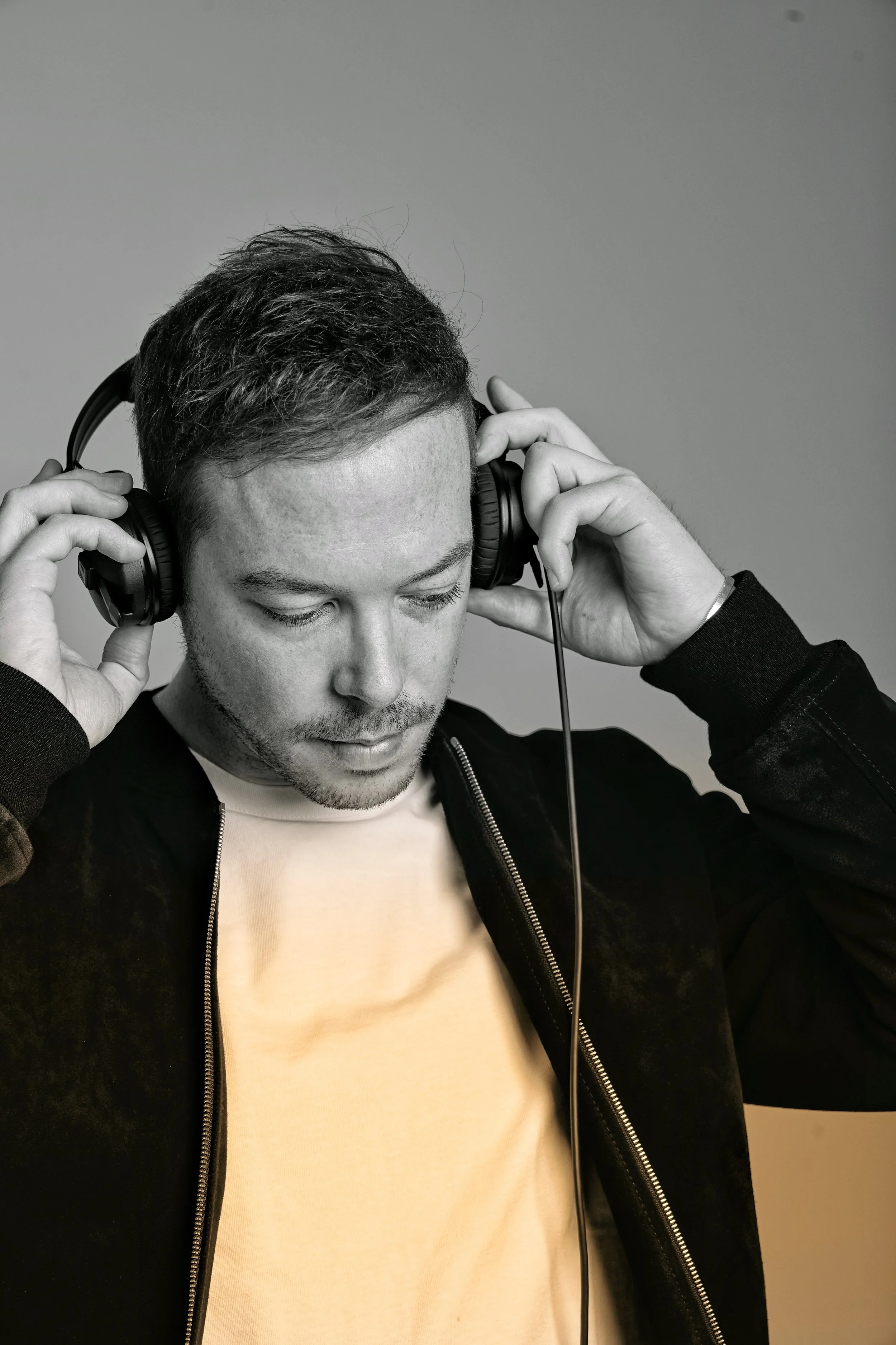 A young man with short dark hair and a beard is wearing large over-ear headphones and a black zip-up jacket, looking down with his eyes closed.
