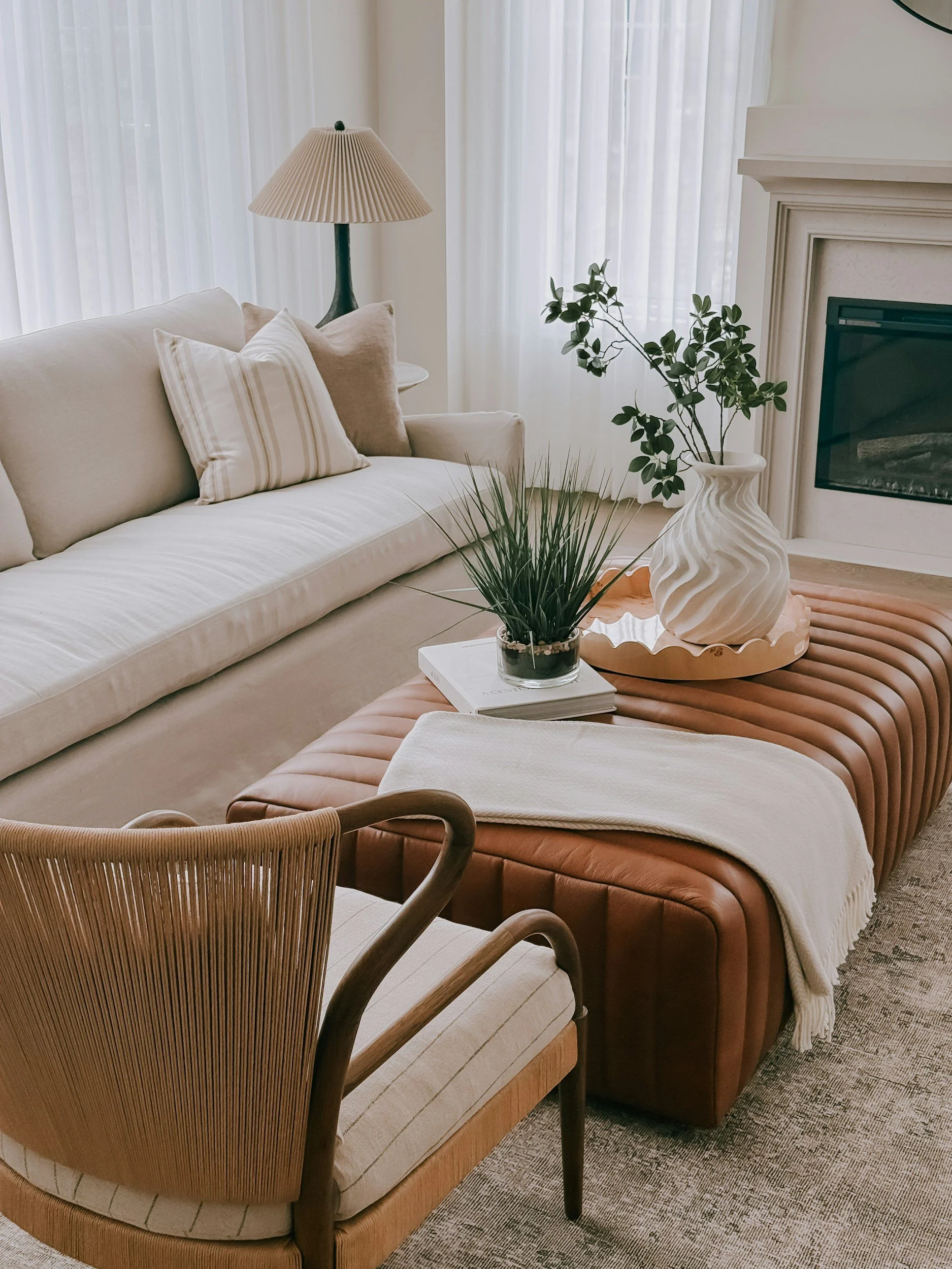 A living room with beige sofa, striped and plain pillows, a glass-top coffee table with a white vase and green plant, a brown leather ottoman, a side lamp, and white curtains.
