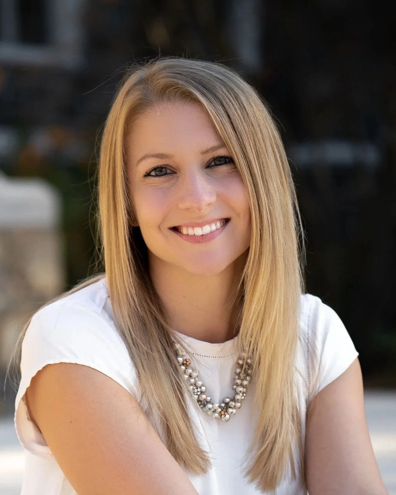 A young woman with long, blonde hair smiling at the camera, wearing a white top and a pearl necklace, outdoors with blurred background.