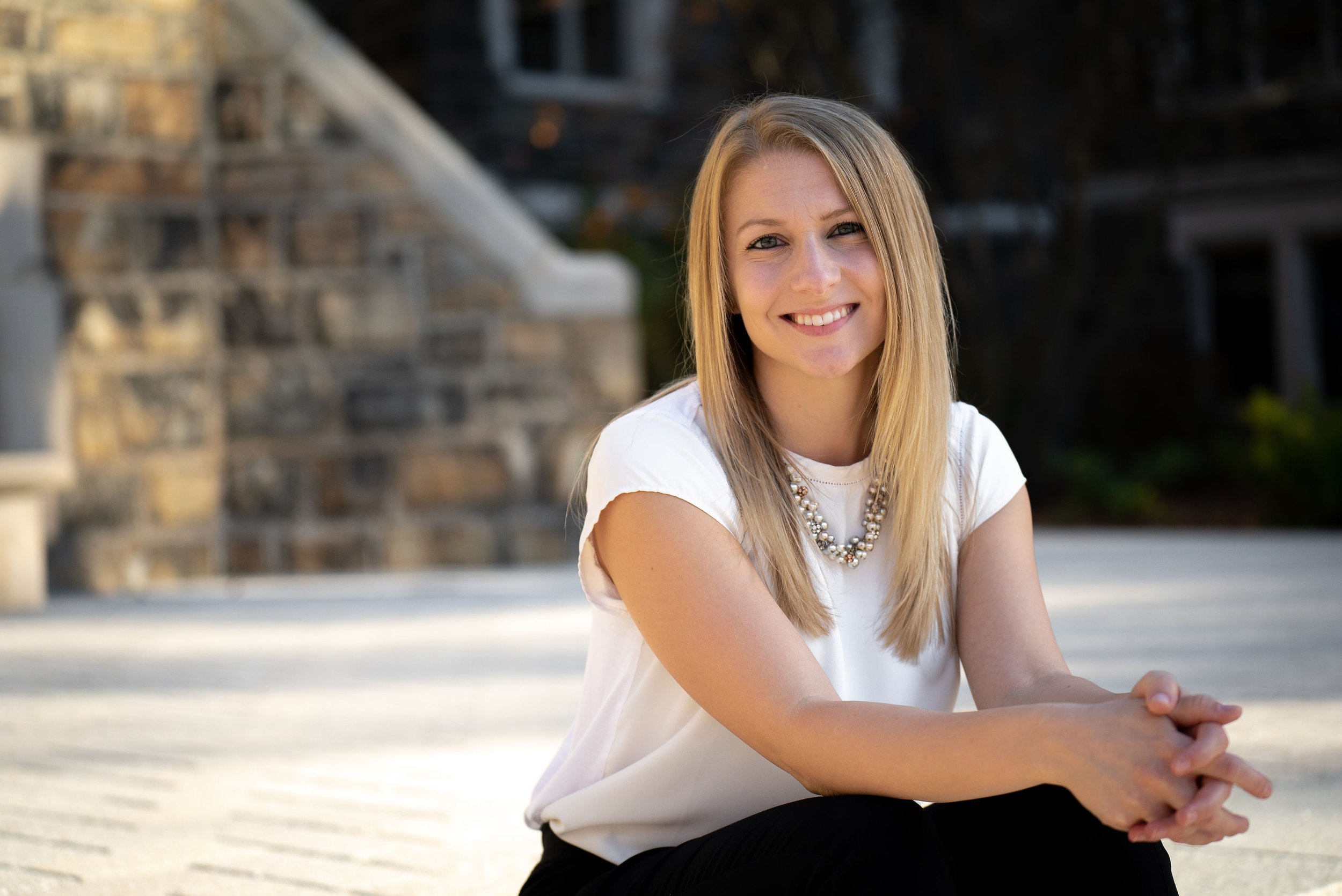 A young woman with long blonde hair, smiling, wearing a white top and a layered pearl necklace, sitting outdoors with a blurred stone building in the background.