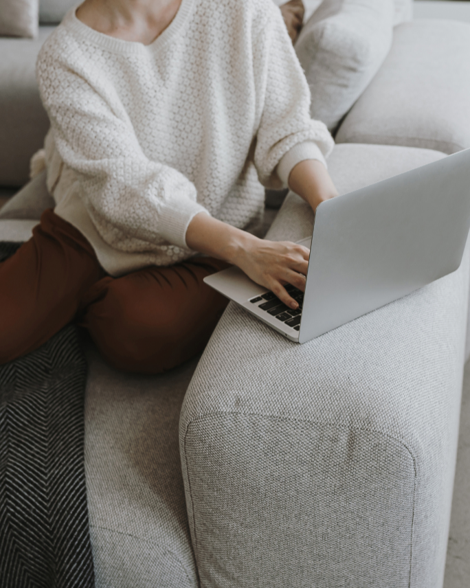 Person sitting on a beige sofa using a silver laptop, wearing a white knitted sweater and brown pants.