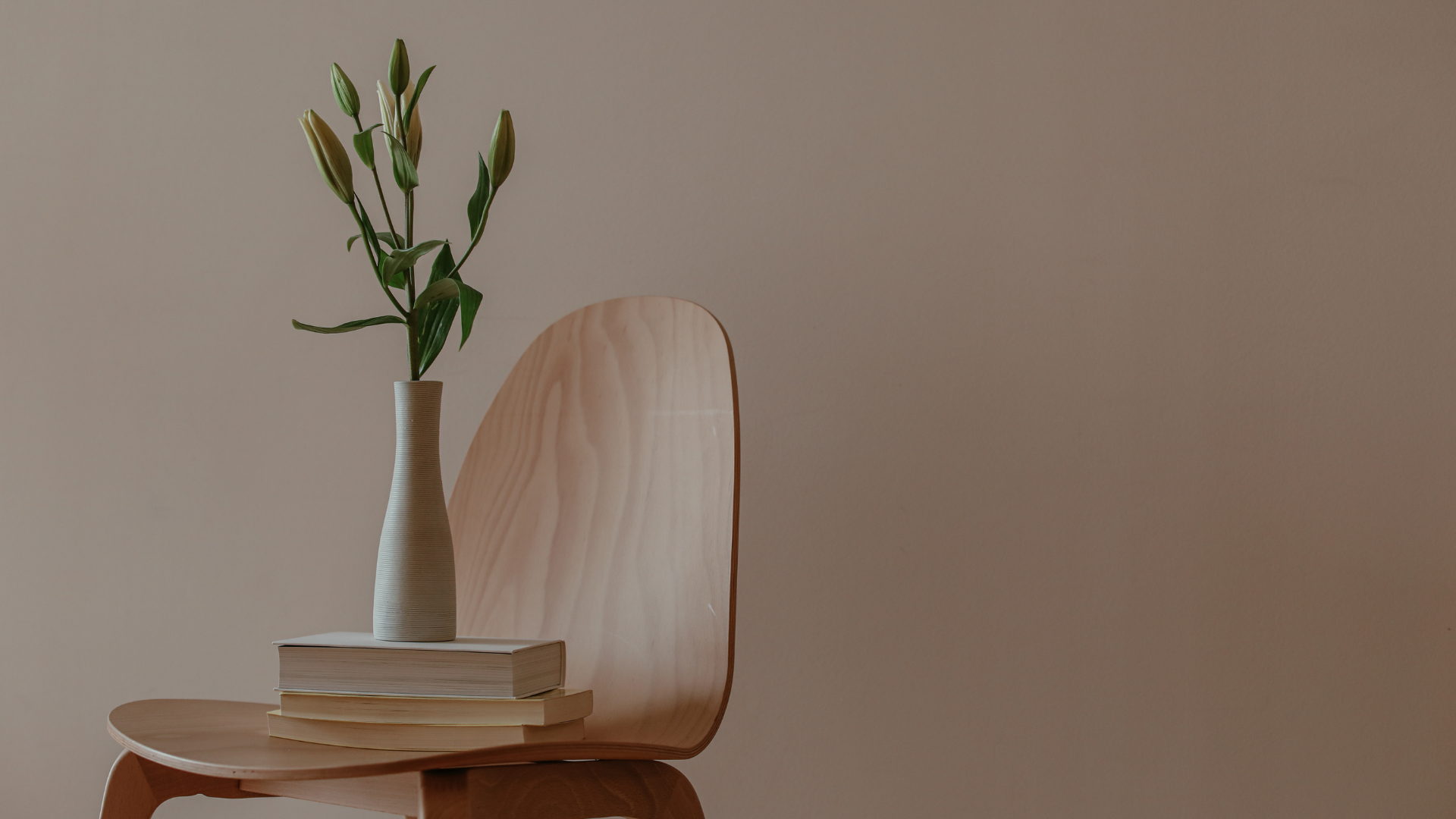 A minimalist decor with a wooden chair, a stack of books, a tall white vase with floral details, and a large wooden panel on the wall.