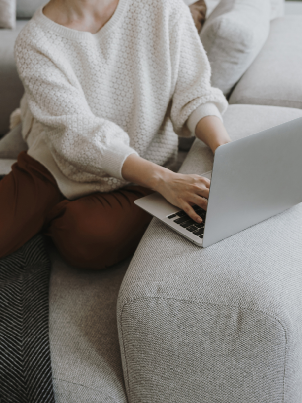 A person wearing a cream-colored sweater sitting on a sofa and working on a laptop.
