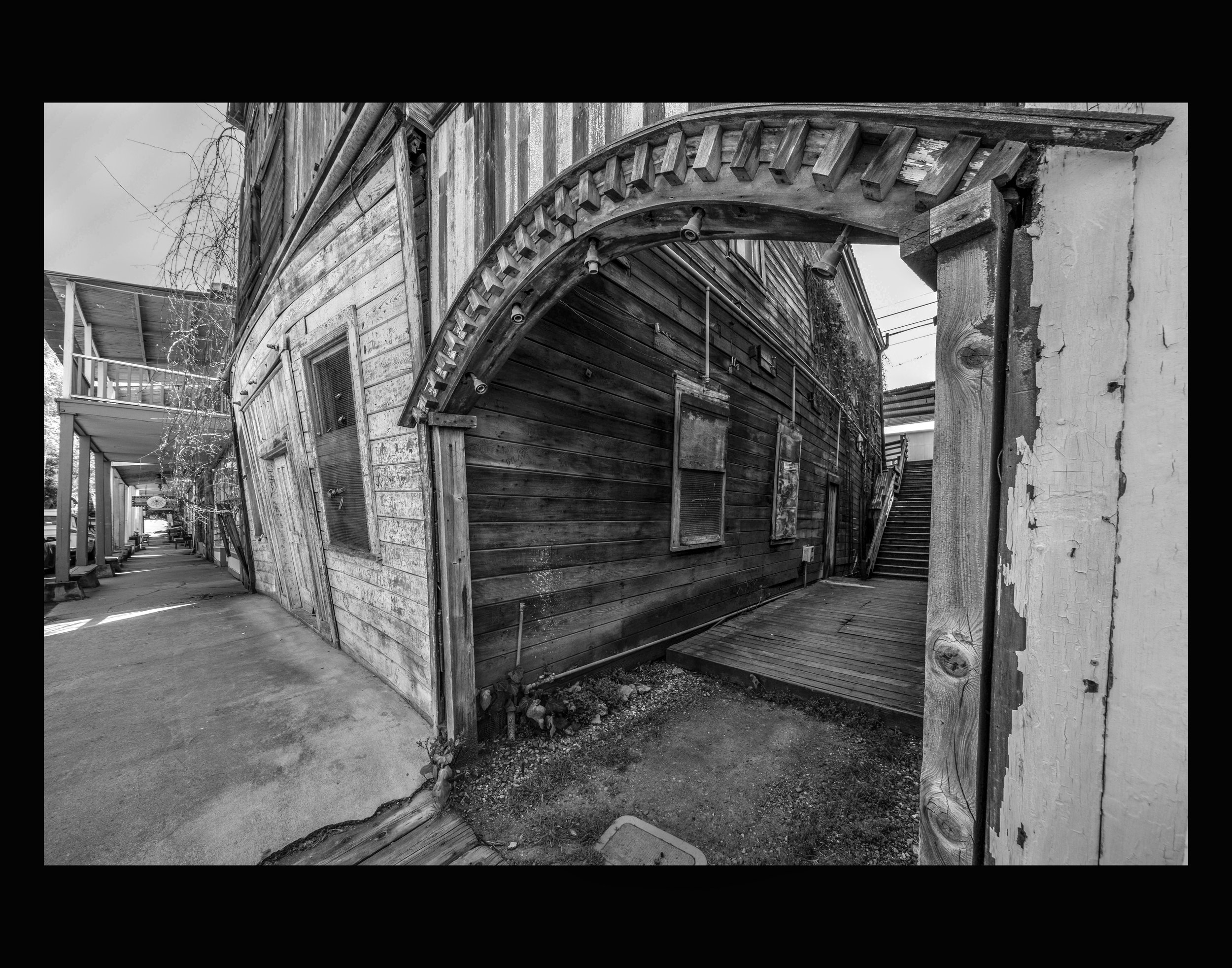 Black and white photograph of a weathered wooden building with an arched entryway, partially damaged and leaning to one side, with visible cracks in the wall, and electric wires overhead.
