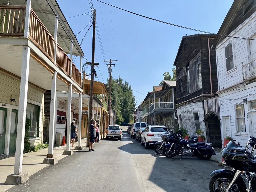 A narrow street lined with old wooden buildings, some with balconies, parked cars and motorcycles on the side, and a few people standing on the sidewalk under a clear blue sky.