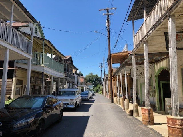 A street scene in a small town with cars parked along both sides and wooden buildings with balconies, some with businesses, under a clear blue sky.