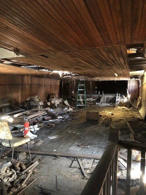 Interior of the "third" floor with a damaged ceiling and debris scattered across the floor, including chairs, boxes, box springs and construction materials.