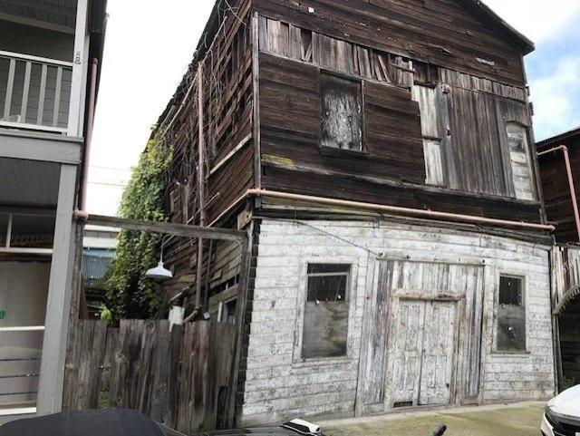 A two-story building with a weathered, dark wooden upper level and a white washed wood facade on the lower level, showing signs of age and disrepair.