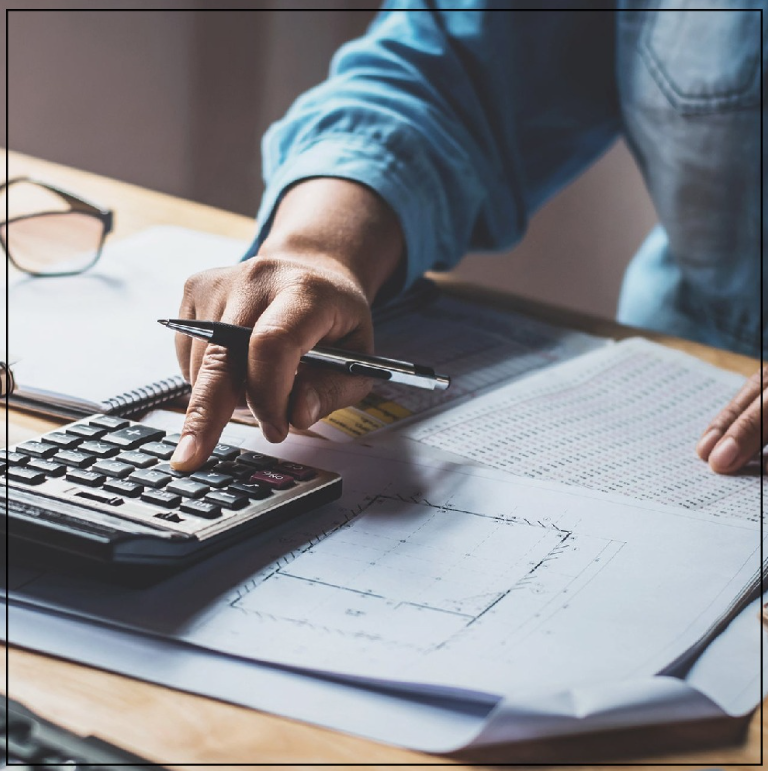 A person's hand pressing buttons on a calculator, with papers, a notebook, and a pen on a wooden desk.