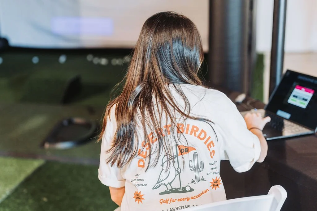 Back of a person with long brown hair, wearing a white t-shirt with orange and black graphics and text, sitting at a table and using a laptop in an indoor setting.
