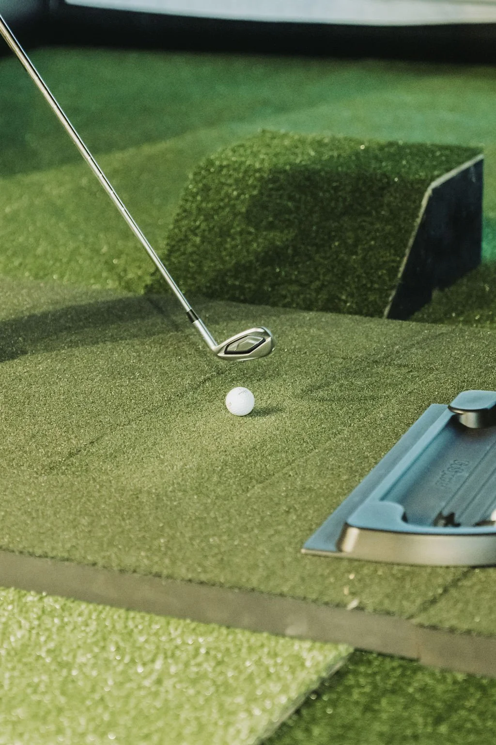 A golf ball on an artificial green mat with a golf club positioned to hit it, and a piece of artificial turf in the background.