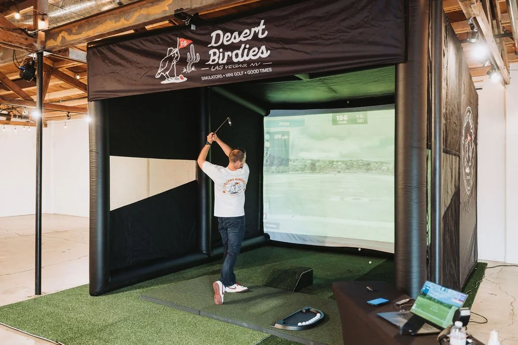 An indoor golf simulator setup titled "Desert Birdies" with a man practicing his golf swing inside a black inflatable enclosure. The enclosure has a graphic of a desert scene and is labeled as being in Las Vegas, Nevada, offering simulators, mini gol